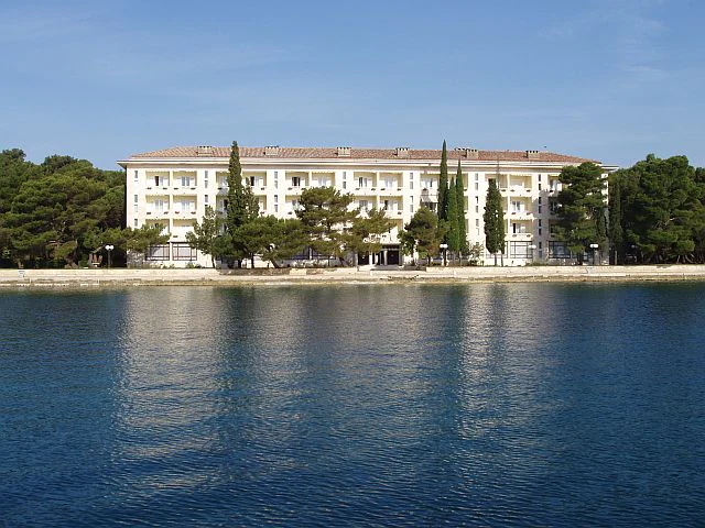 White multi-story hotel building with red roof and trees, situated on a shoreline with calm blue water
