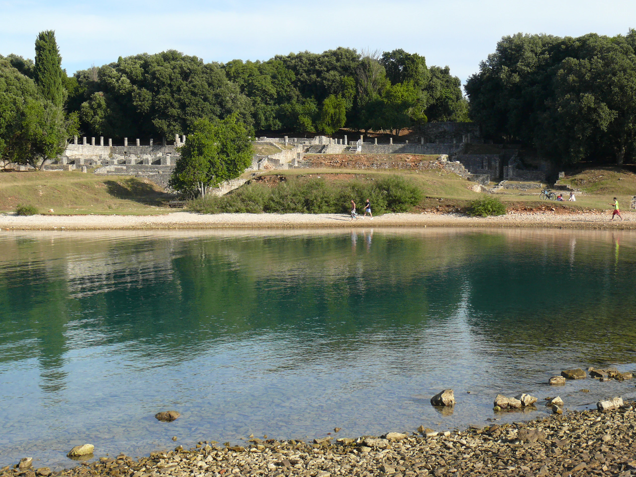 Roman villa ruins along a coastal shore with calm blue water, pebbles, and green trees in the background