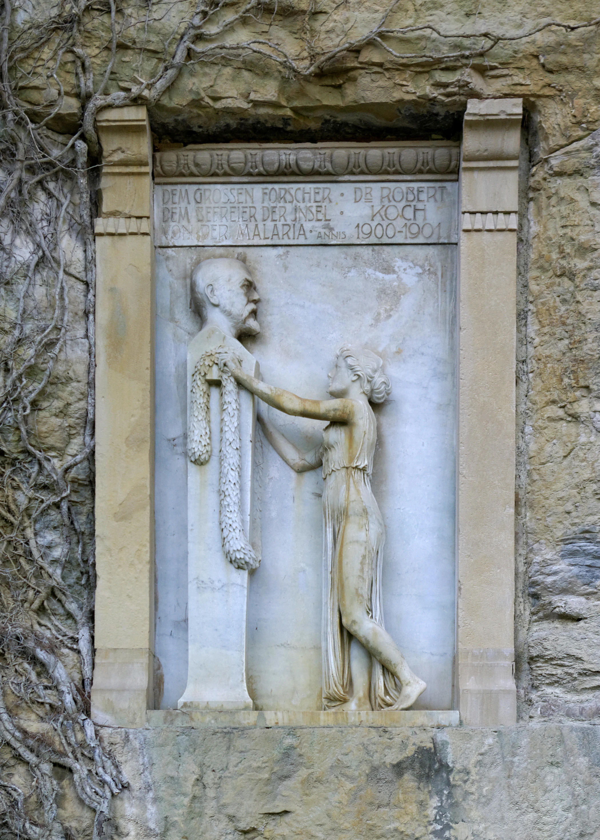 Stone relief sculpture showing a standing male figure and a female figure touching his shoulder, with inscribed text above, mounted on a stone wall with vines