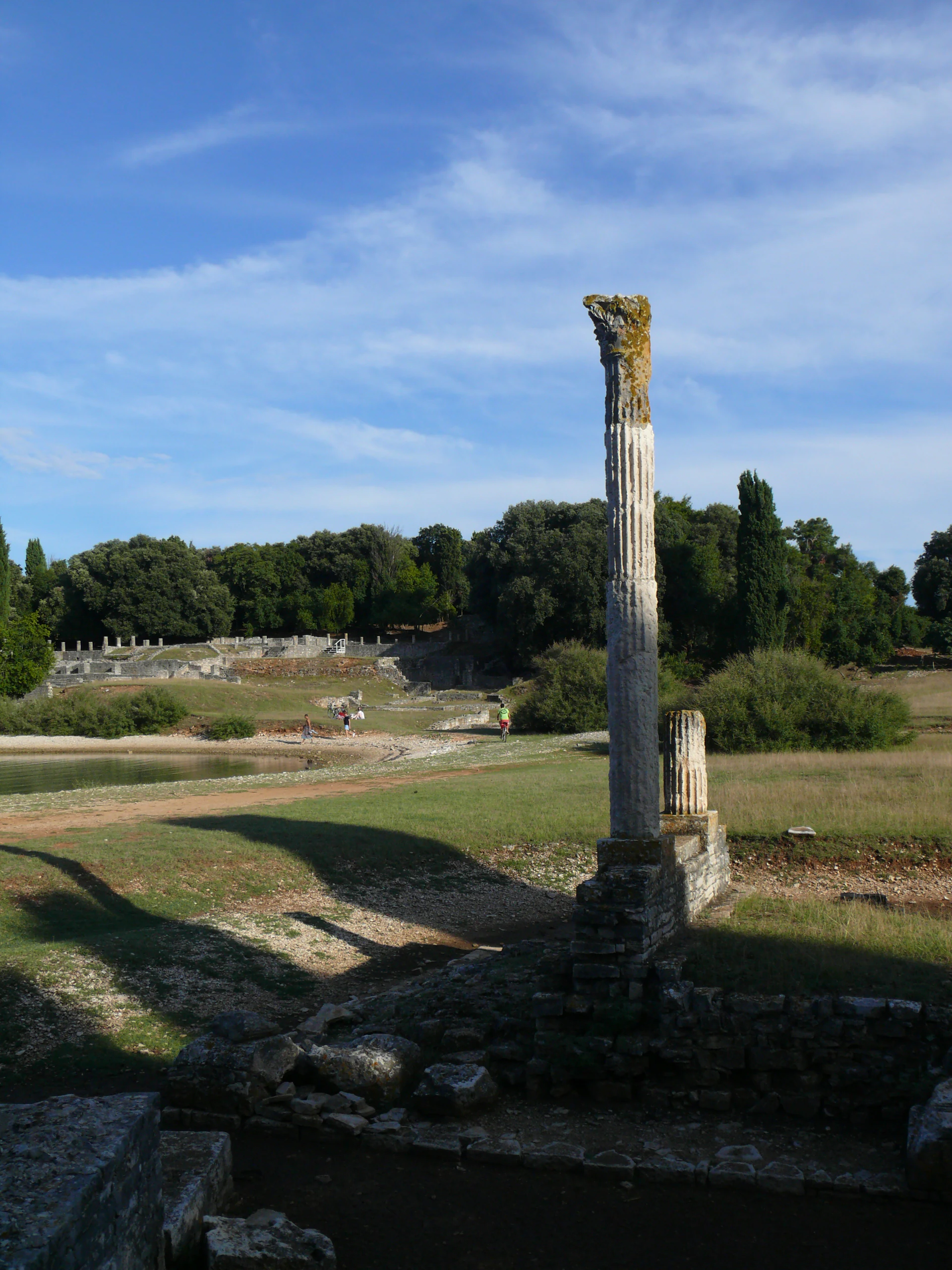 Tall weathered stone column standing amidst grassy field with ancient stone ruins, trees, and clear blue sky