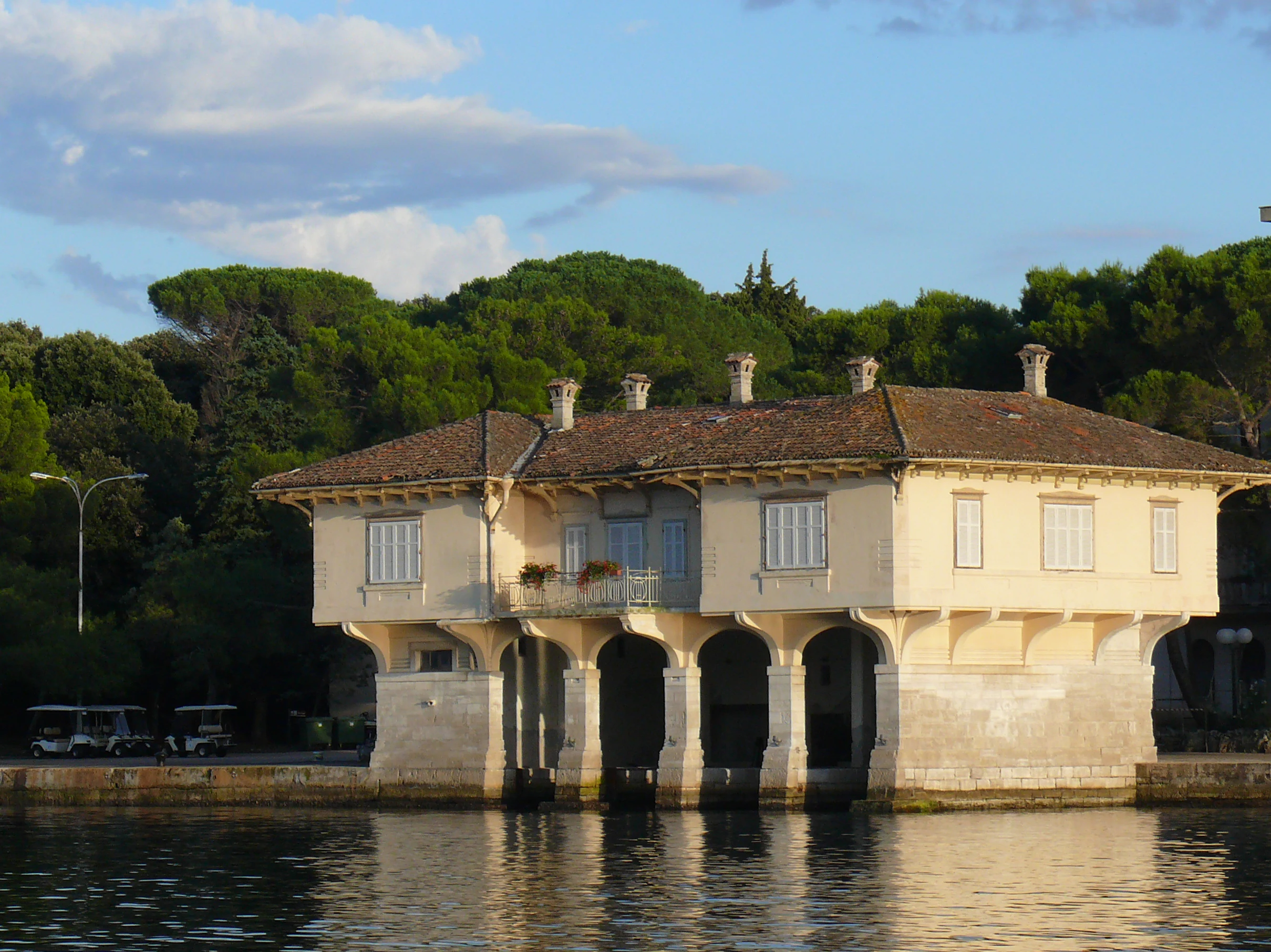 A beige villa with a red-tiled roof and arched openings standing on stilts over calm water, with trees in the background.