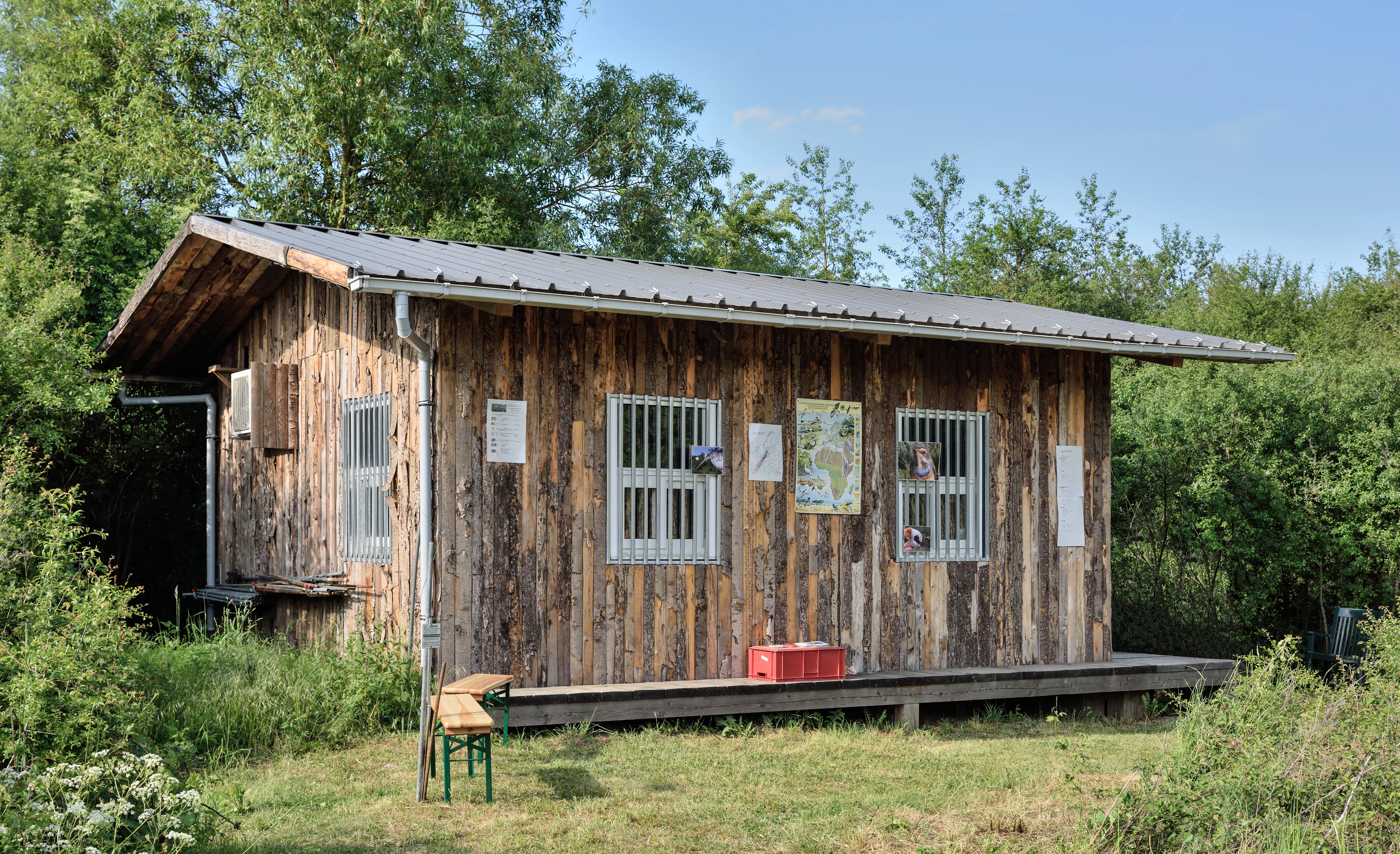 A rustic wooden cabin with a metal roof surrounded by trees and green vegetation at the Brill nature reserve, featuring barred windows and informative signs on the exterior walls.