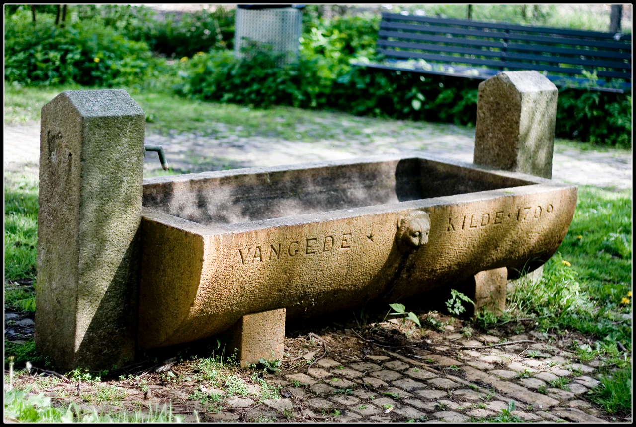 A historic stone water trough or spring fountain with the text Vangede Kilde 1709 carved into the side, situated on a stone paved path with a park bench in the background.