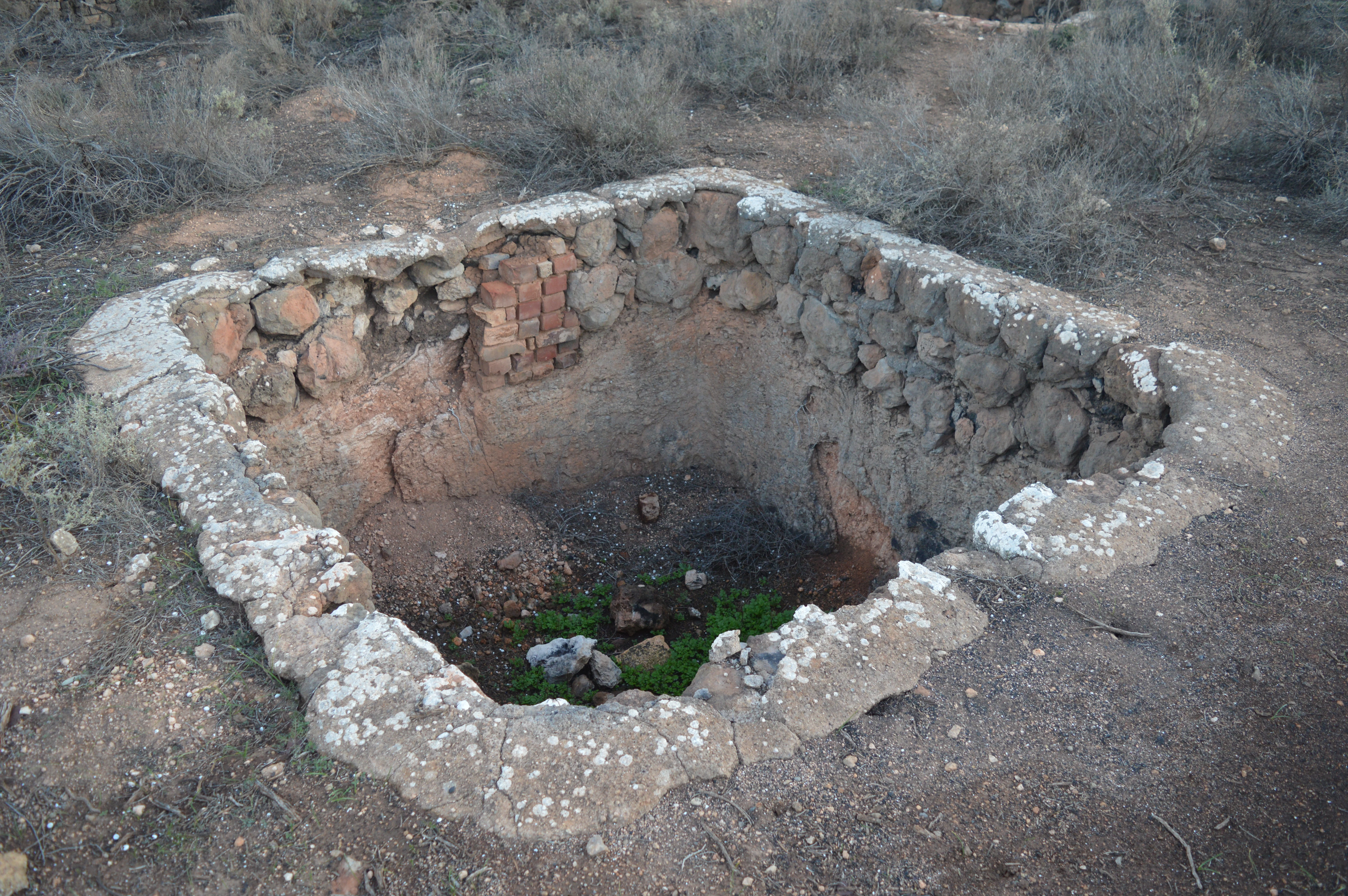 A square, stone and brick lined pit built into the ground, showing signs of abandonment and age, surrounded by dry, arid landscape vegetation.