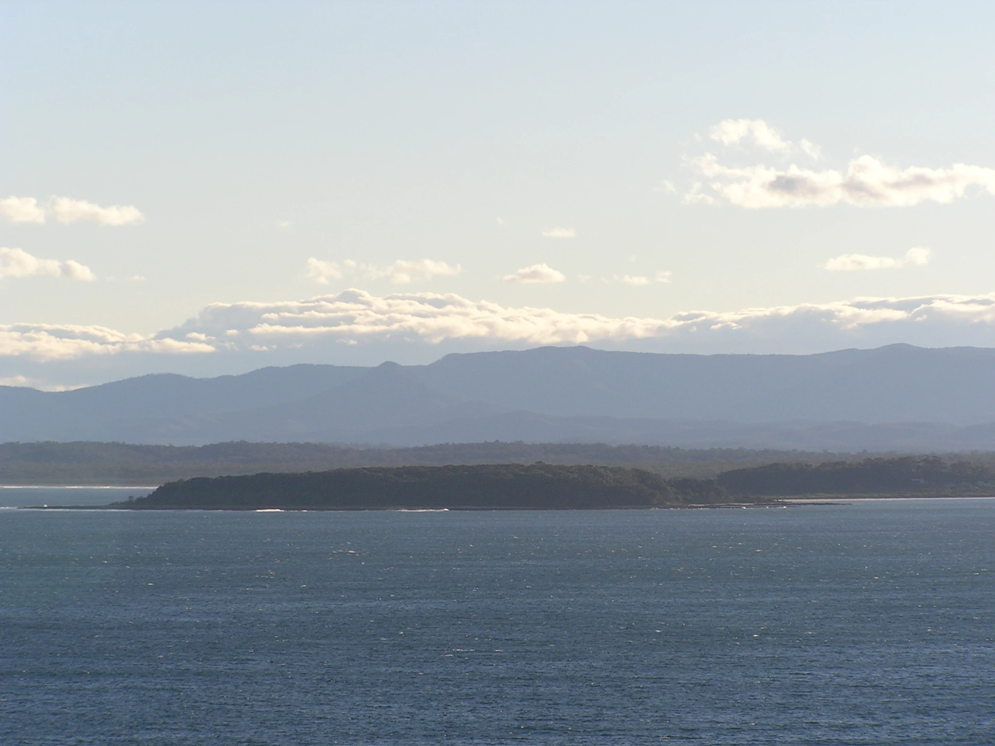 A wide coastal view of Broulee Island appearing as a dark, tree-covered landmass separated from the mainland, with ocean in the foreground and a hazy mountain range in the far background.