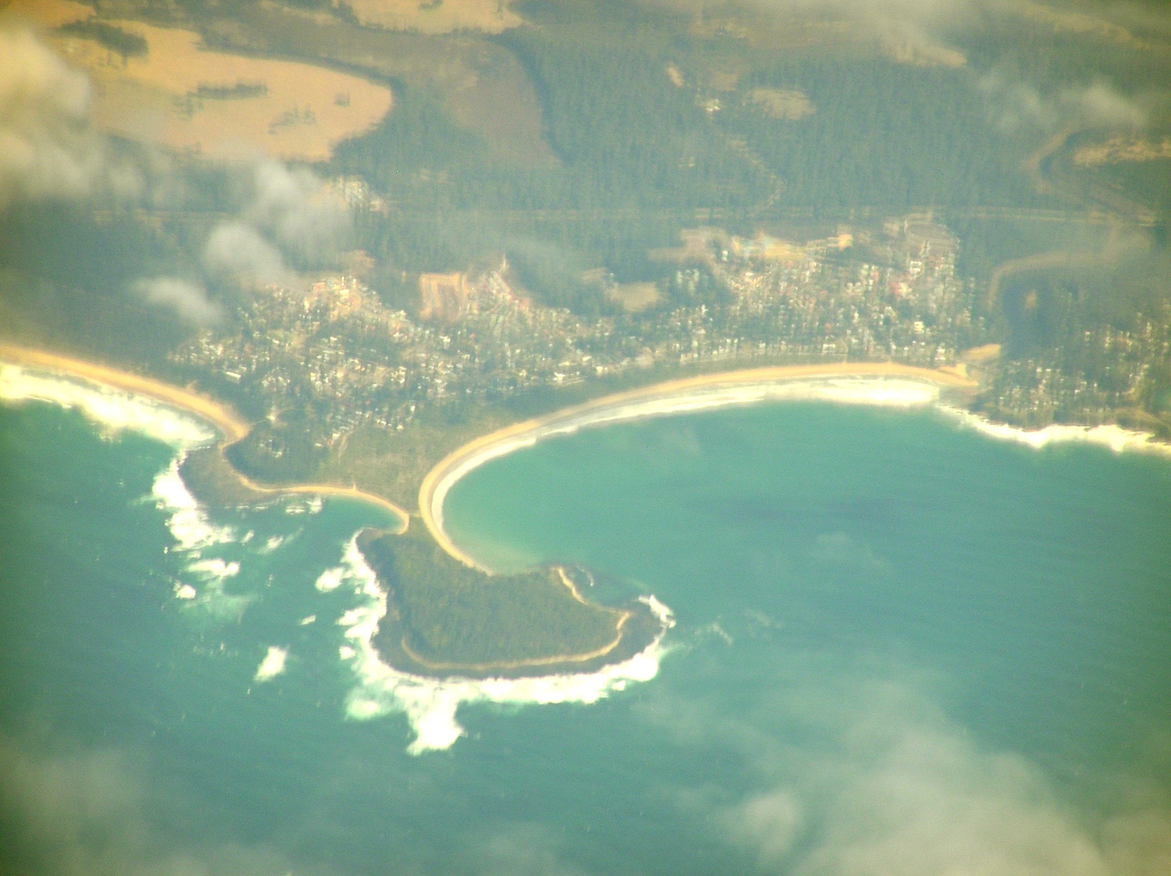 An aerial perspective of Broulee Island, showing the tombolo sandbar connecting the island to the mainland near a coastal town.