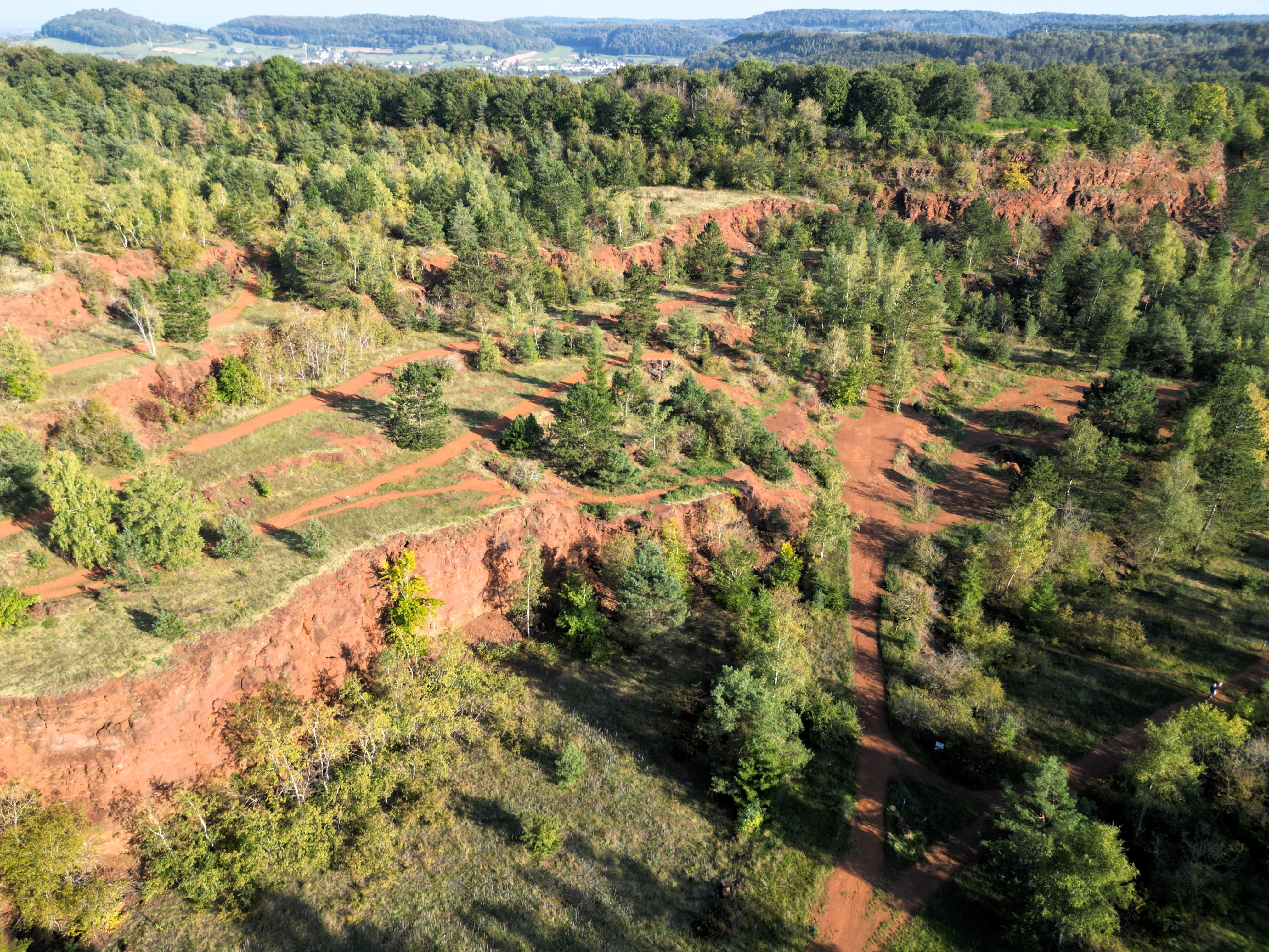 Aerial view of a nature reserve with reddish soil, exposed rock faces, and forest vegetation in a former mining area.