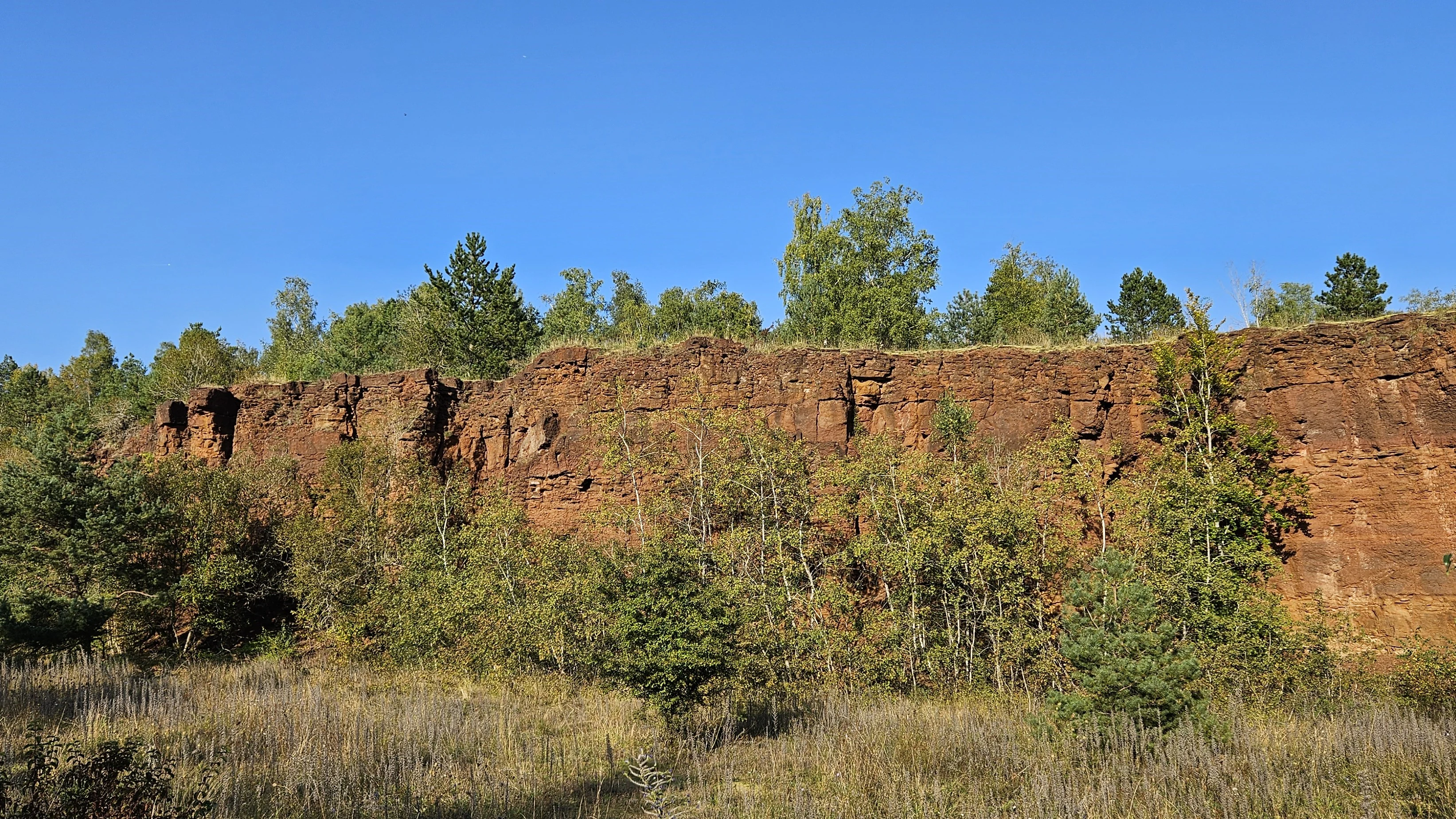 A wide shot of a reddish-brown rock escarpment covered in sparse trees and shrubs, typical of the former mining landscape in Brucherbierg-Lallengerbierg.