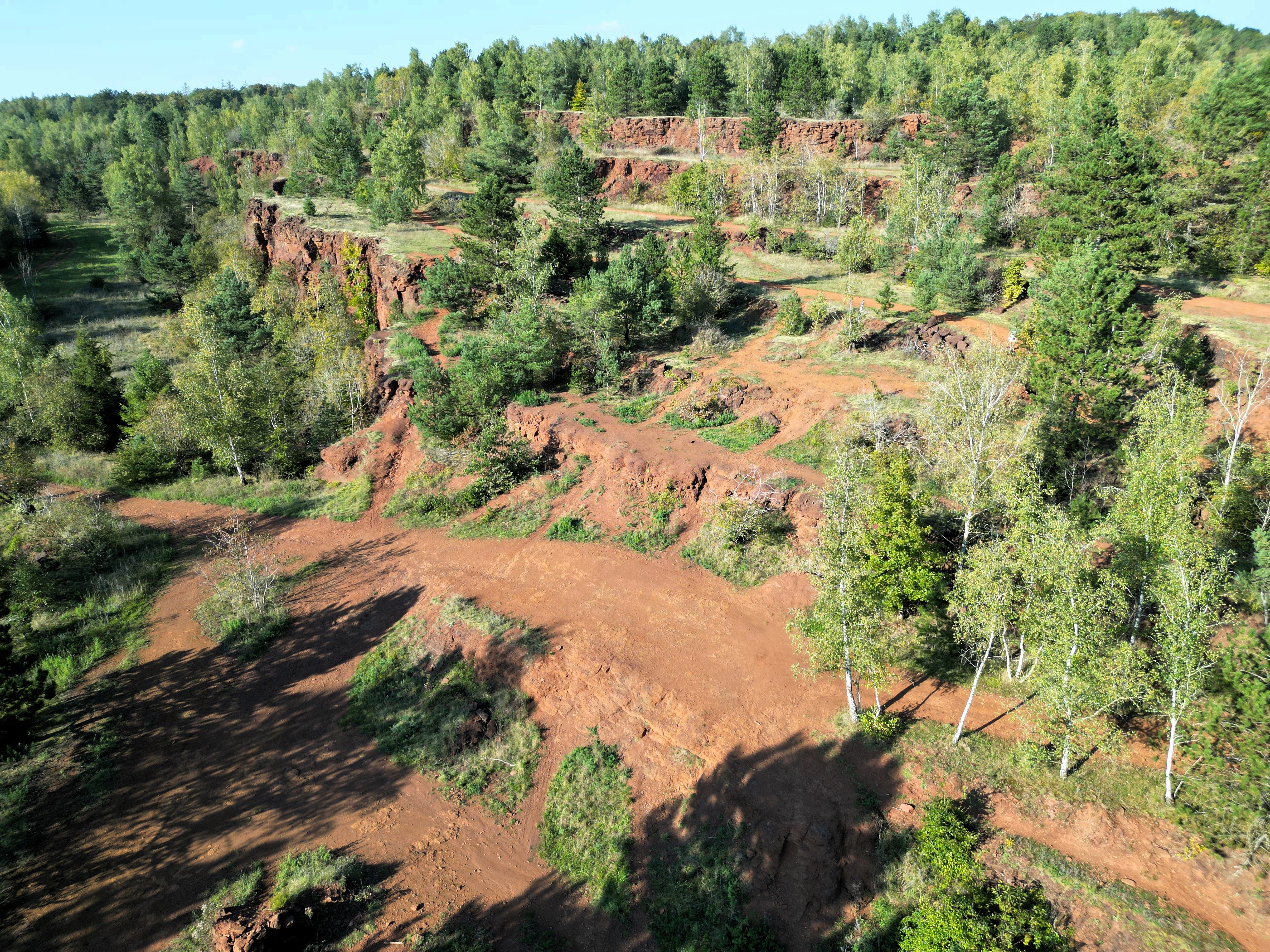 A high-angle view of a former open-cast mining area showing stepped, rust-colored soil terraces surrounded by green trees and shrubs.