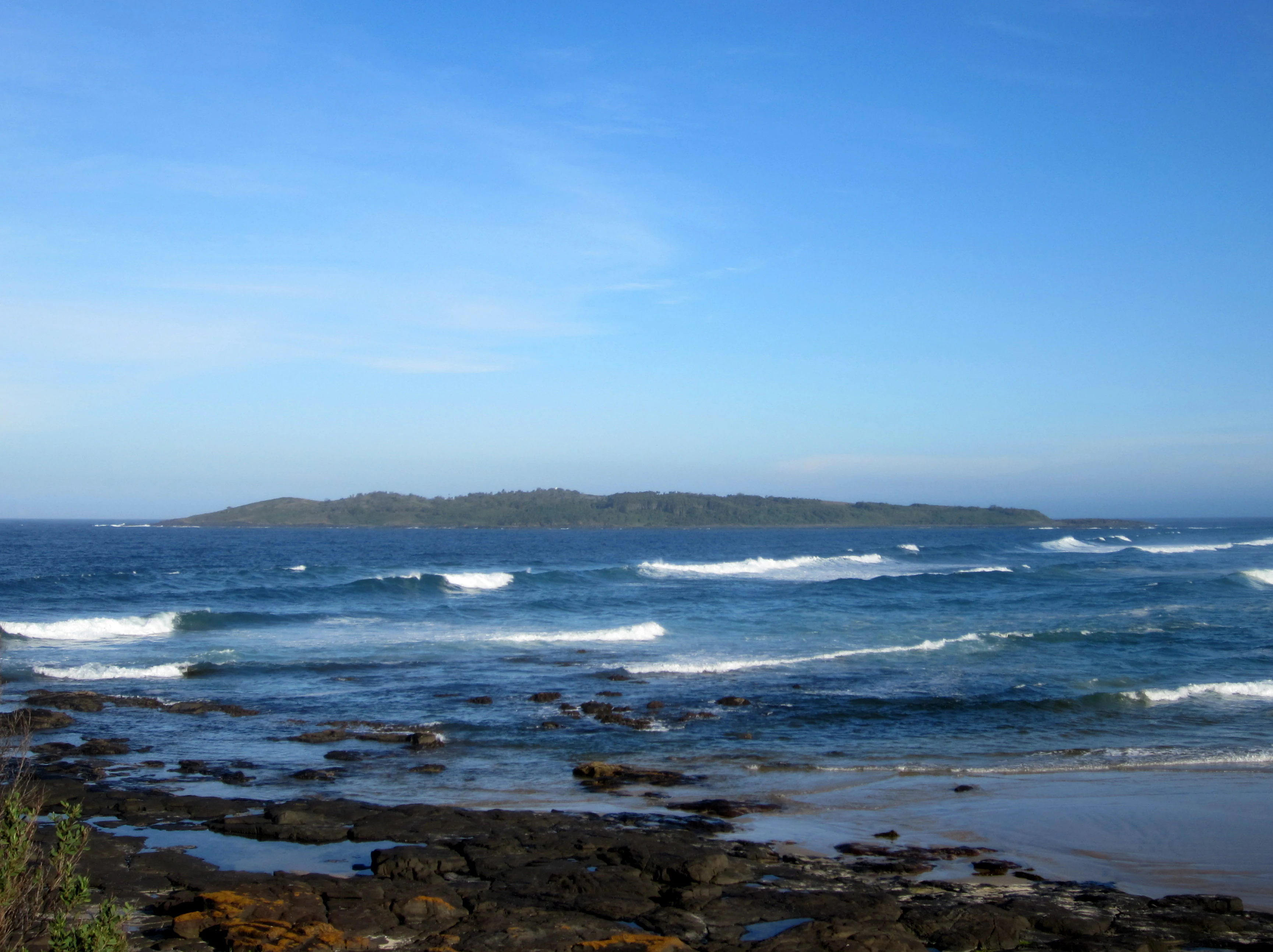 A wide angle view across the blue ocean towards a low lying green island sitting on the horizon under a clear blue sky, with dark rocky foreground and small waves breaking near the shore.