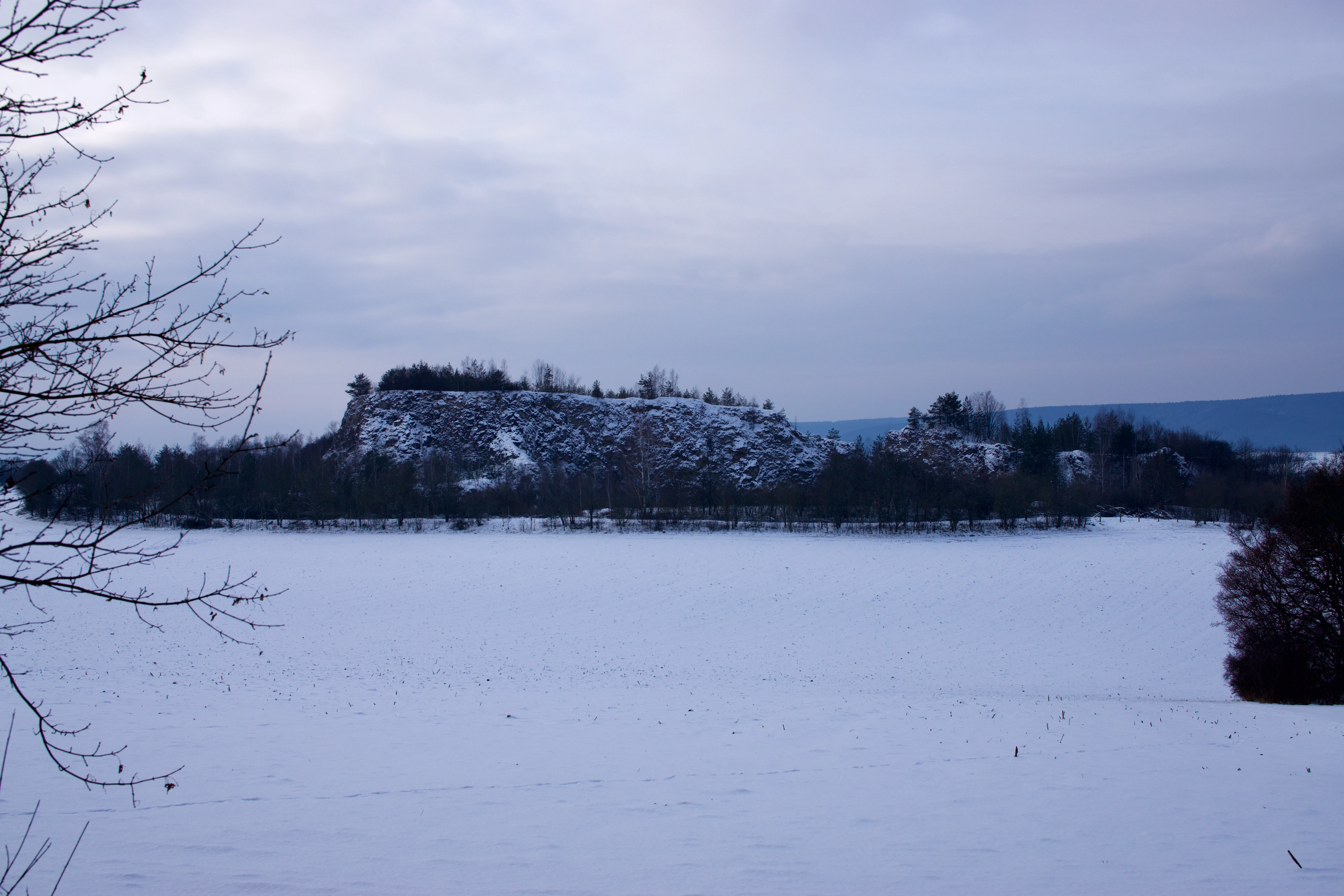 A wide, eye-level landscape shot of a snow-covered hill known as Buchenberg, set against a cloudy winter sky with a snowy field in the foreground.