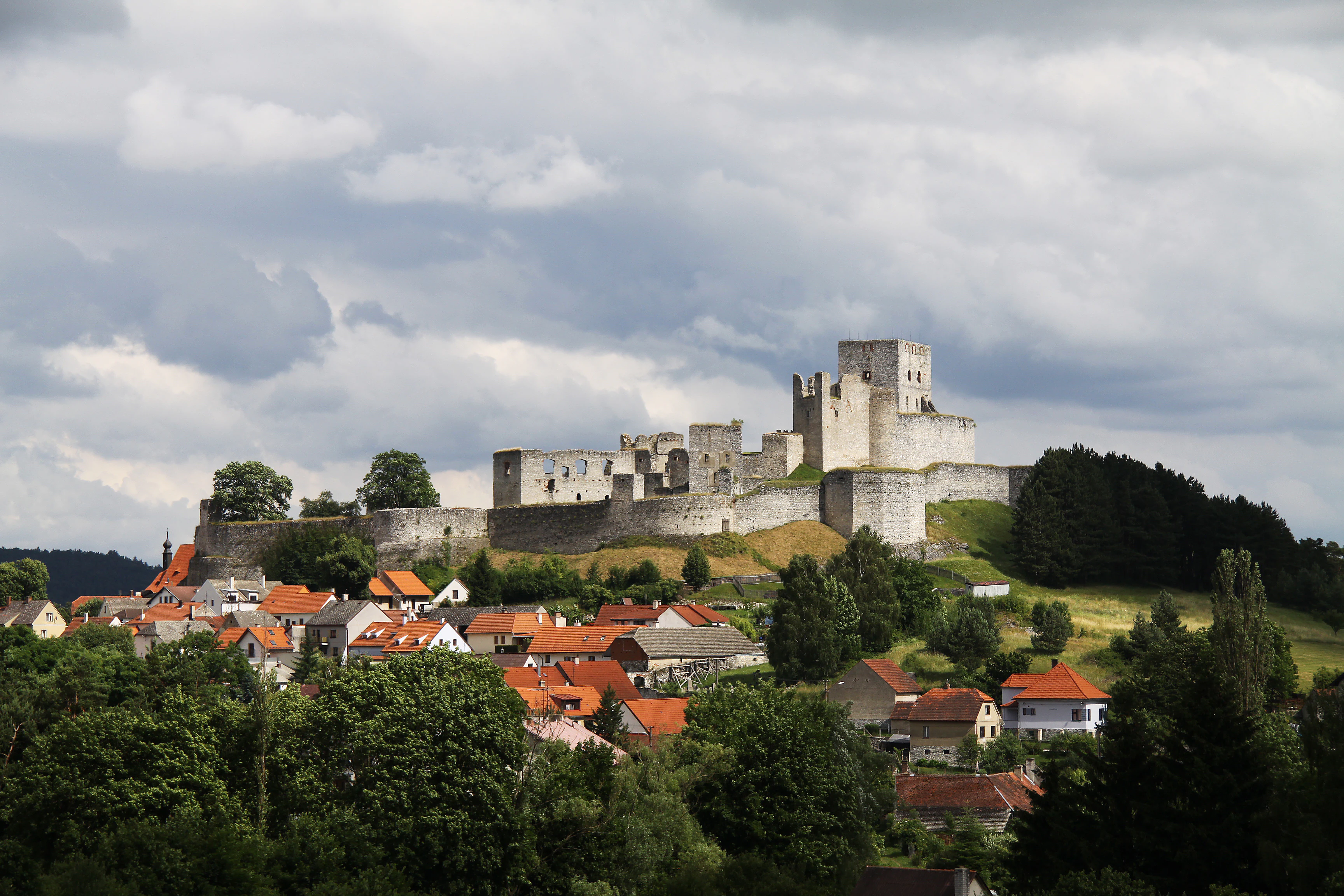 A wide shot showing the extensive stone ruins of Rabi Castle situated on a hilltop above a village with red-roofed houses under a cloudy sky.