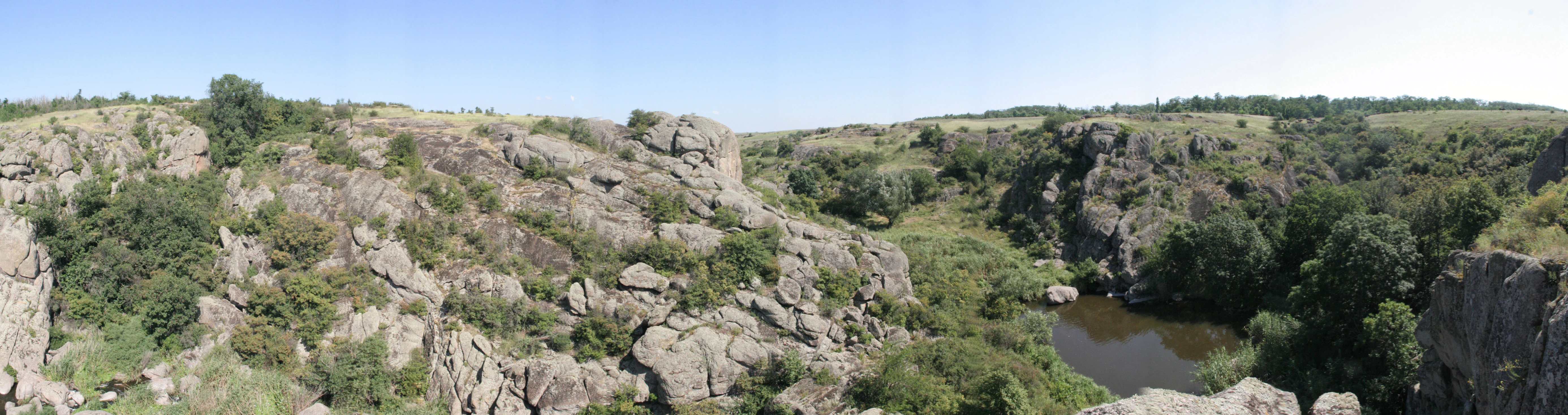 Panoramic view of a rocky canyon with a river flowing through it, surrounded by sparse vegetation and steep rock formations under a clear blue sky