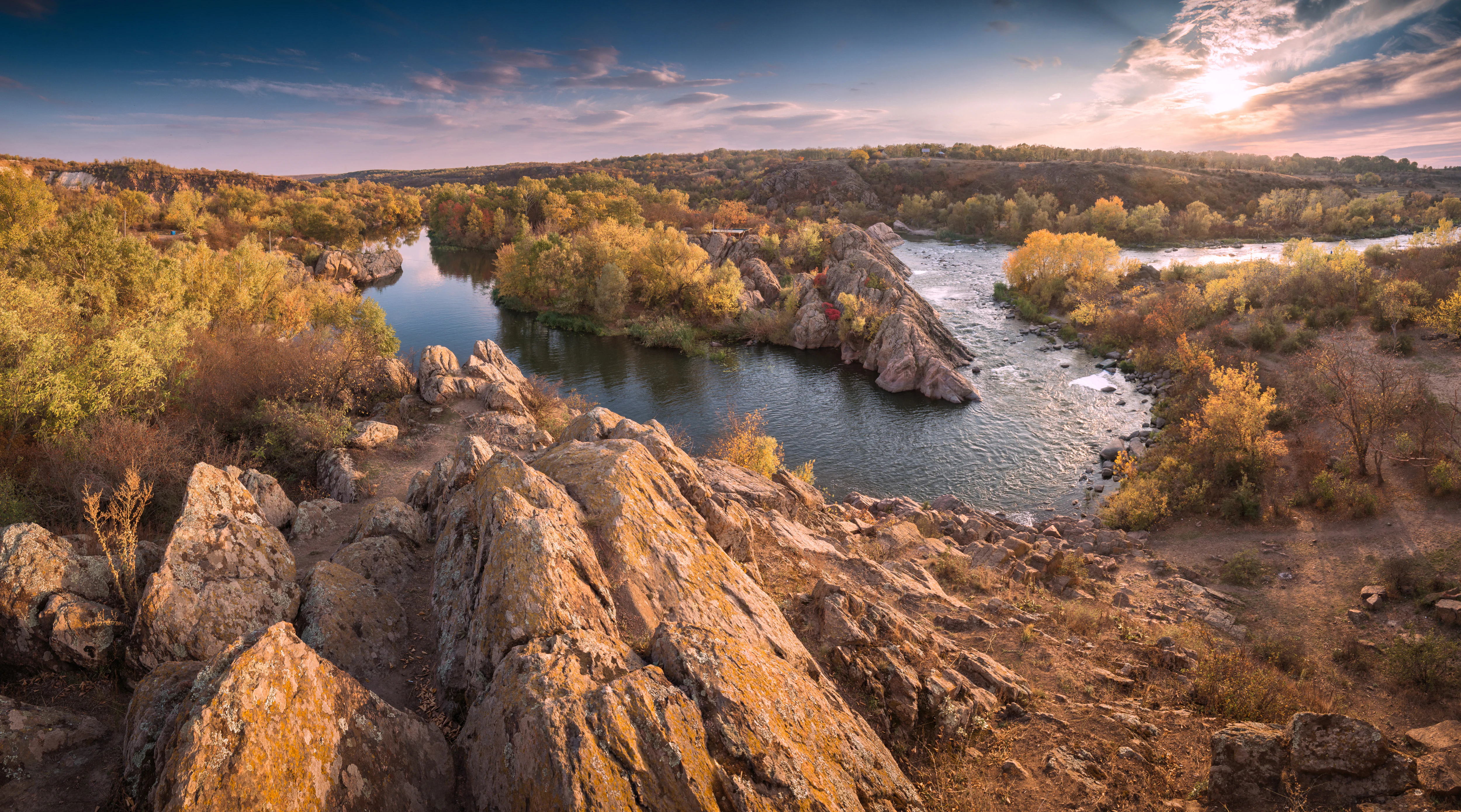 Wide view of a river winding through rocky terrain with autumn-colored trees under a partly cloudy sky