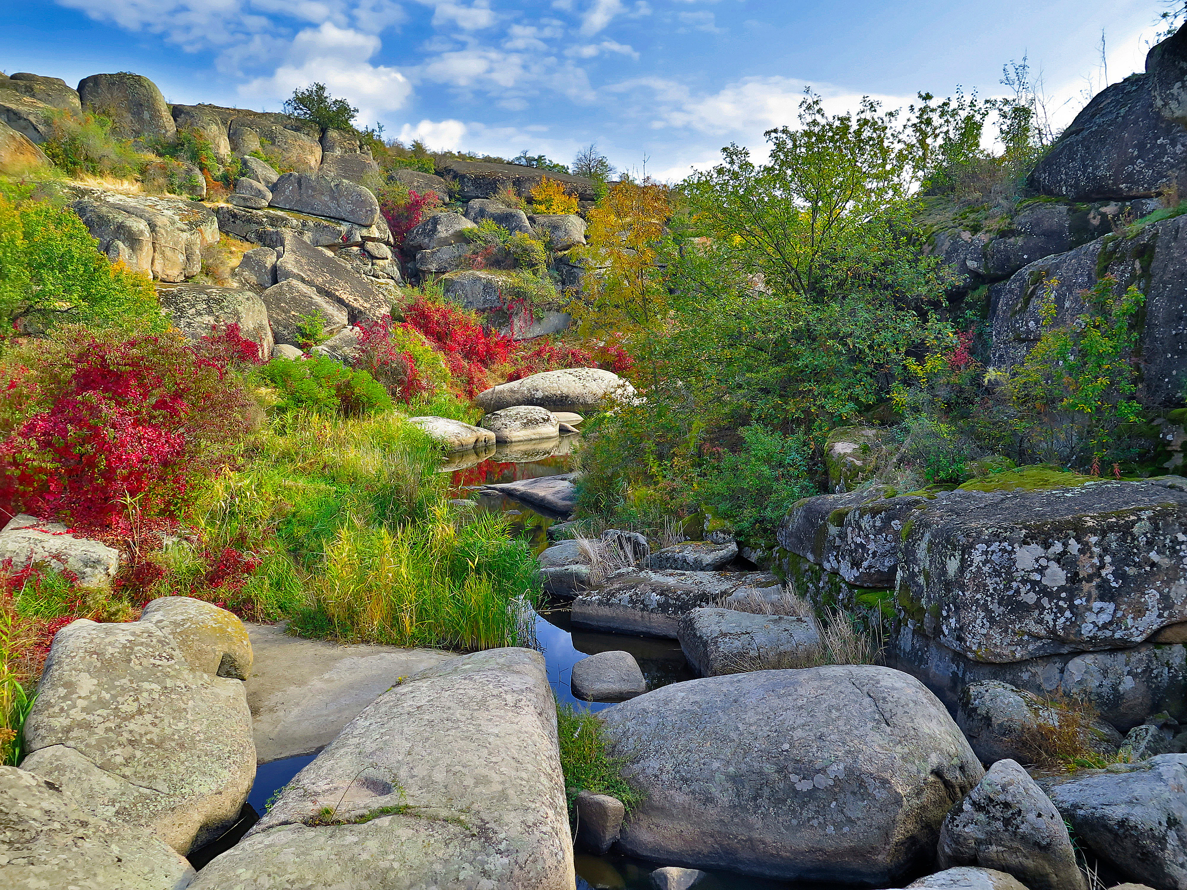 Rocky canyon landscape with small stream, autumn-colored vegetation, and large boulders under partly cloudy sky