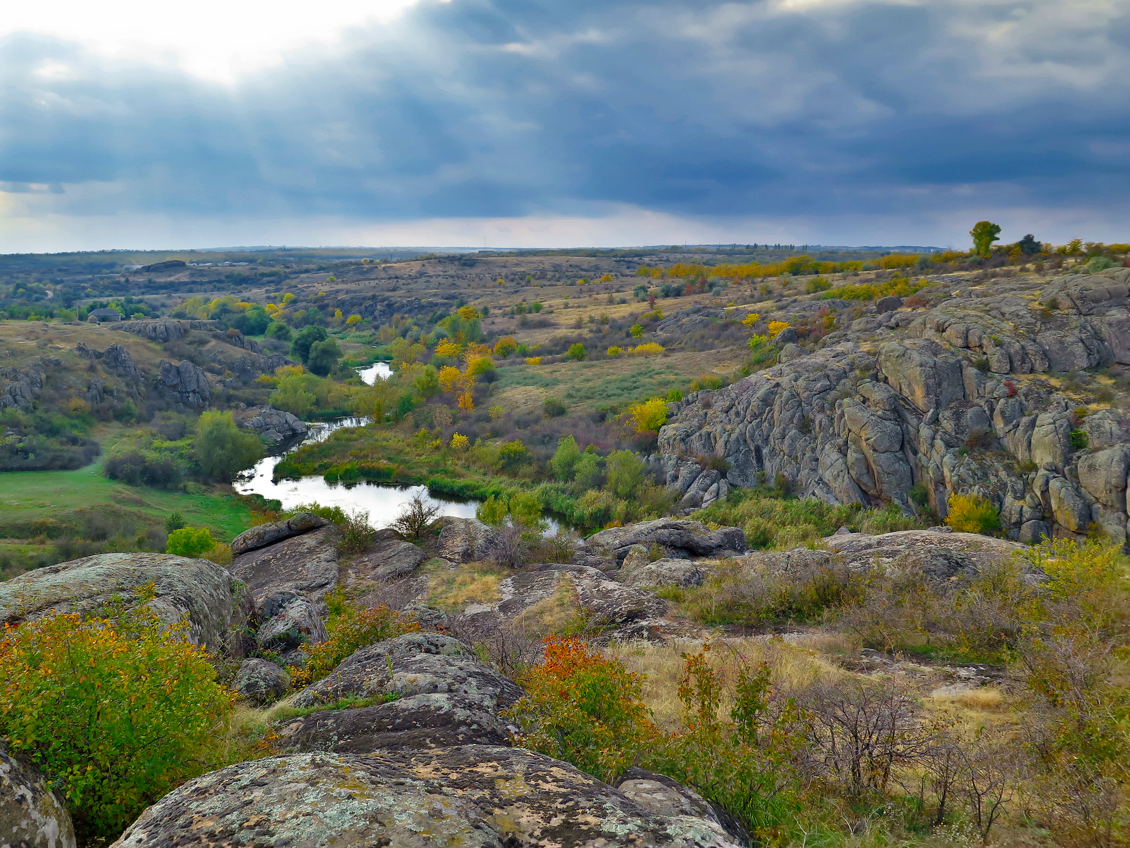 A wide view of a canyon with a winding river, rocky cliffs, and autumn foliage under a cloudy sky