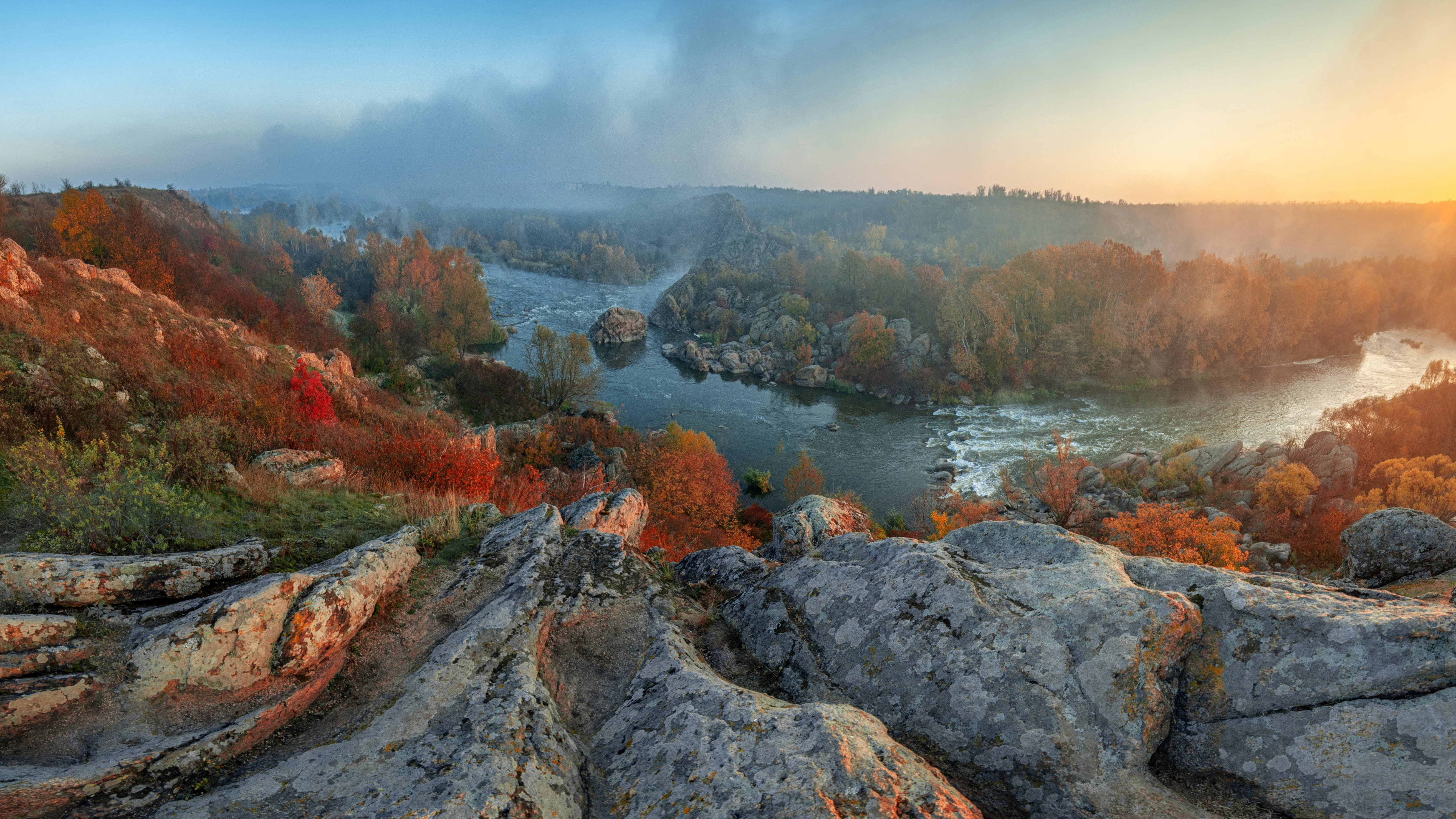 Rocky riverbank with autumn-colored trees and a winding river under a sunset sky