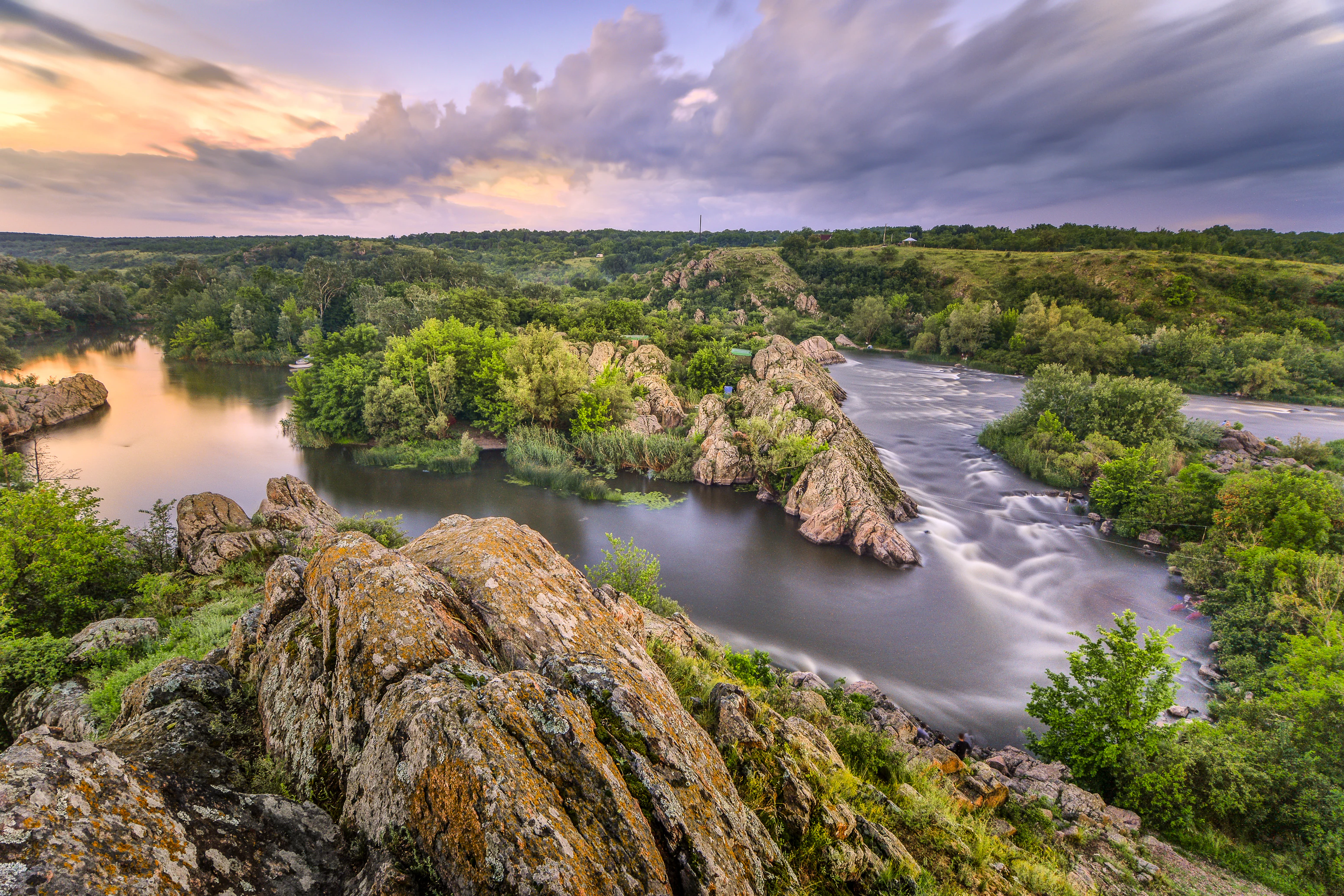 Wide landscape view of the Southern Bug River featuring a prominent bend surrounded by rocky outcrops and lush green vegetation under a partly cloudy evening sky