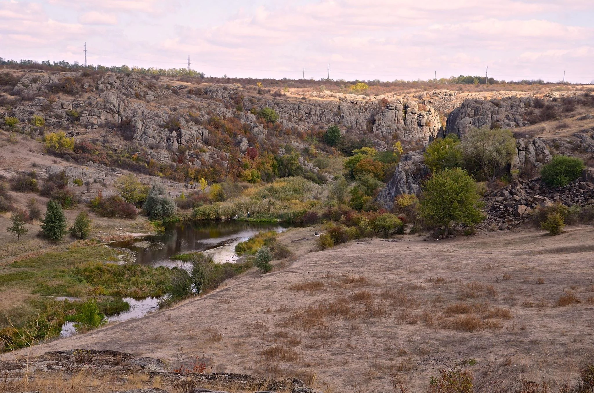 A wide view of a canyon with a river flowing through rocky cliffs, dry grassy terrain, and scattered trees under a partly cloudy sky