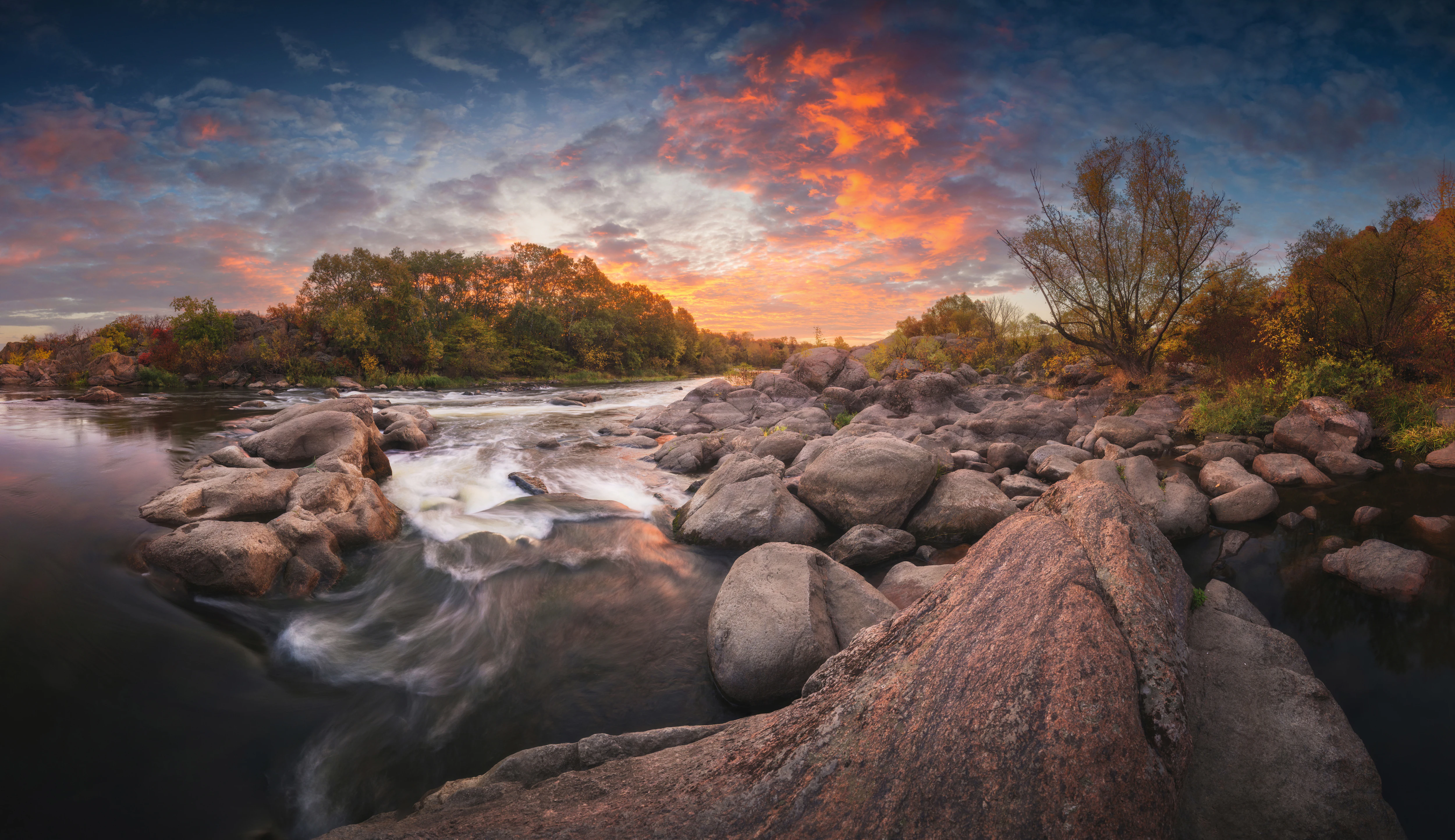 River with rapids flowing over large rocks during dawn, surrounded by trees and a colorful sky with orange and blue clouds.