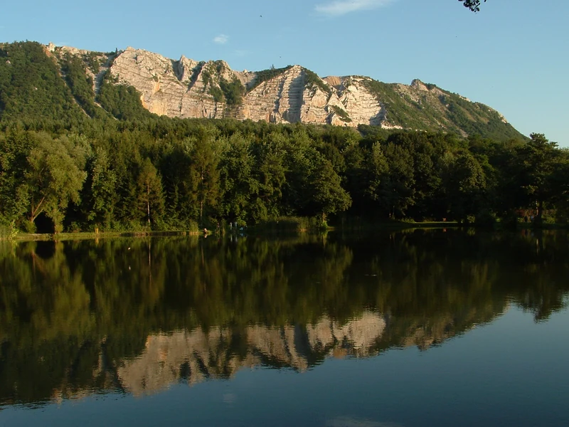 A calm reservoir reflecting a mountain with rocky cliffs and forested slopes under a clear blue sky