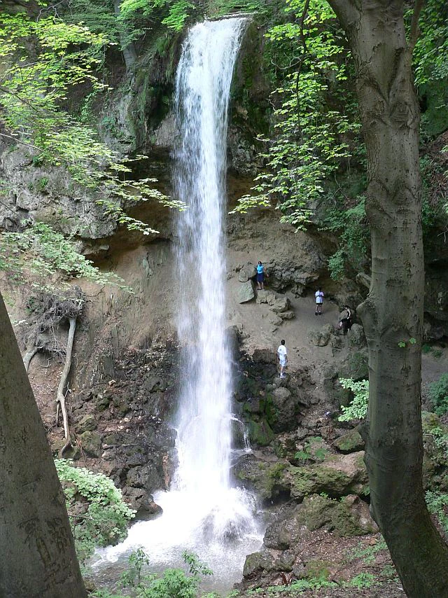 Tall waterfall cascading down a rocky cliff face surrounded by dense forest, with several people visible on the cliffside