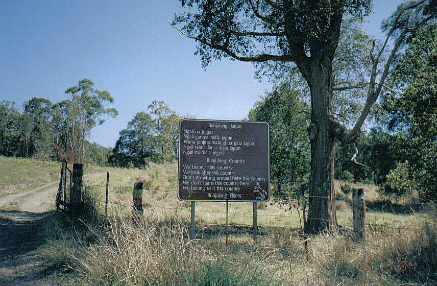 Brown signboard with text in Bundjalung language, standing on grassy field with trees and dirt path in background