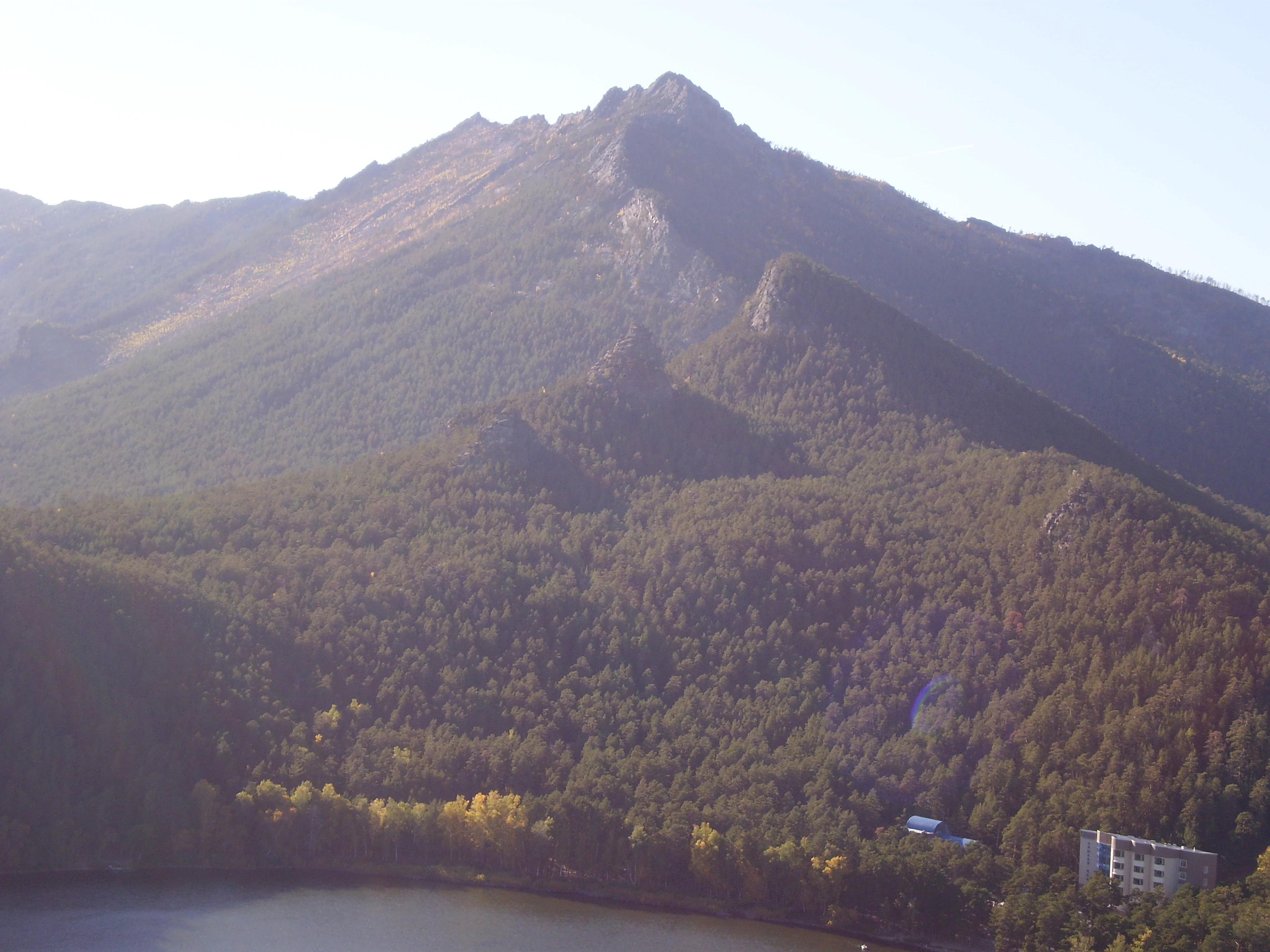 Wide view of a mountainous landscape with dense forests, a lake in the foreground, and distant peaks under clear skies