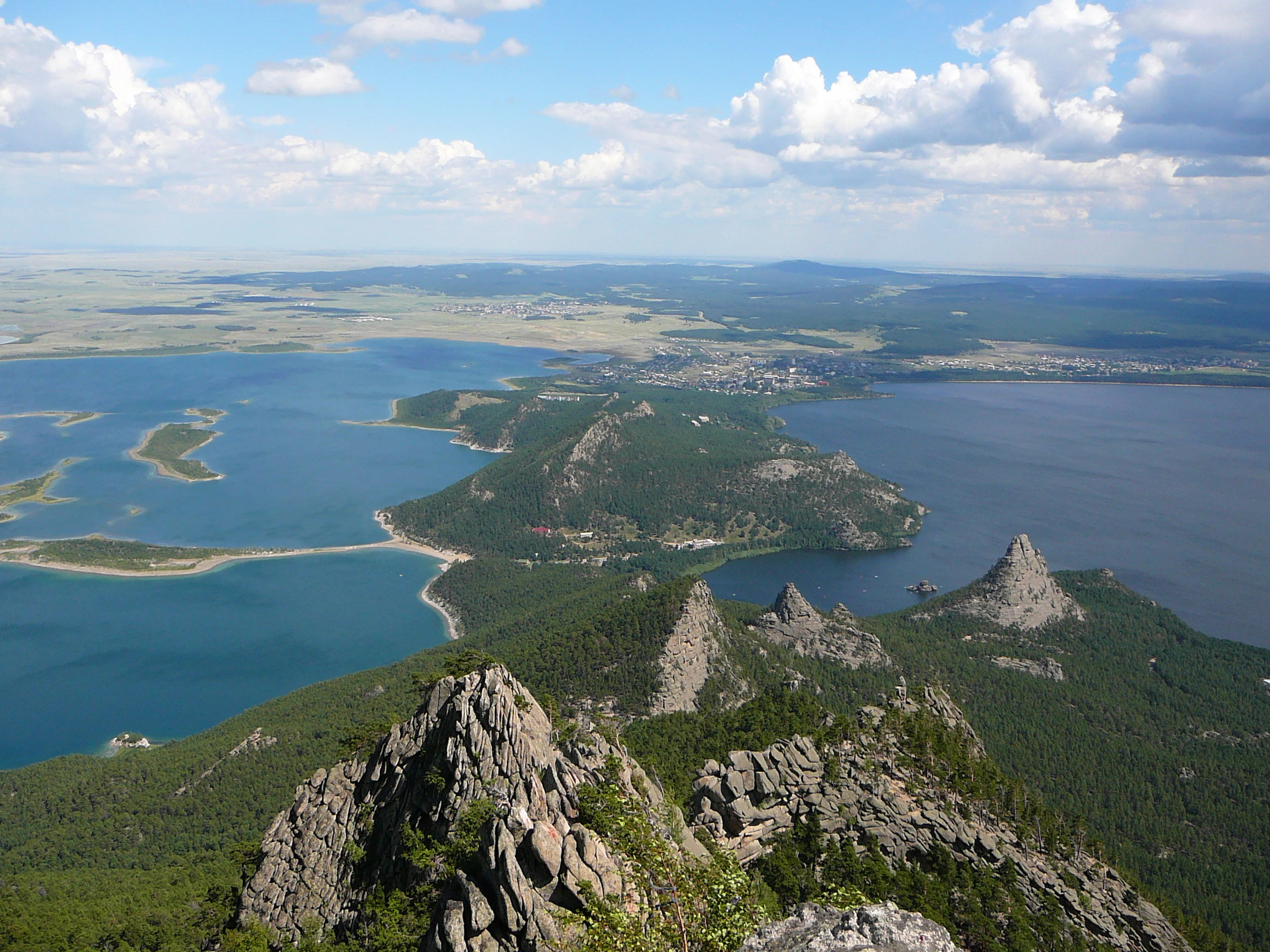 Panoramic landscape view showing two large lakes connected by a narrow isthmus, surrounded by rocky mountain peaks and dense forest under a partly cloudy sky