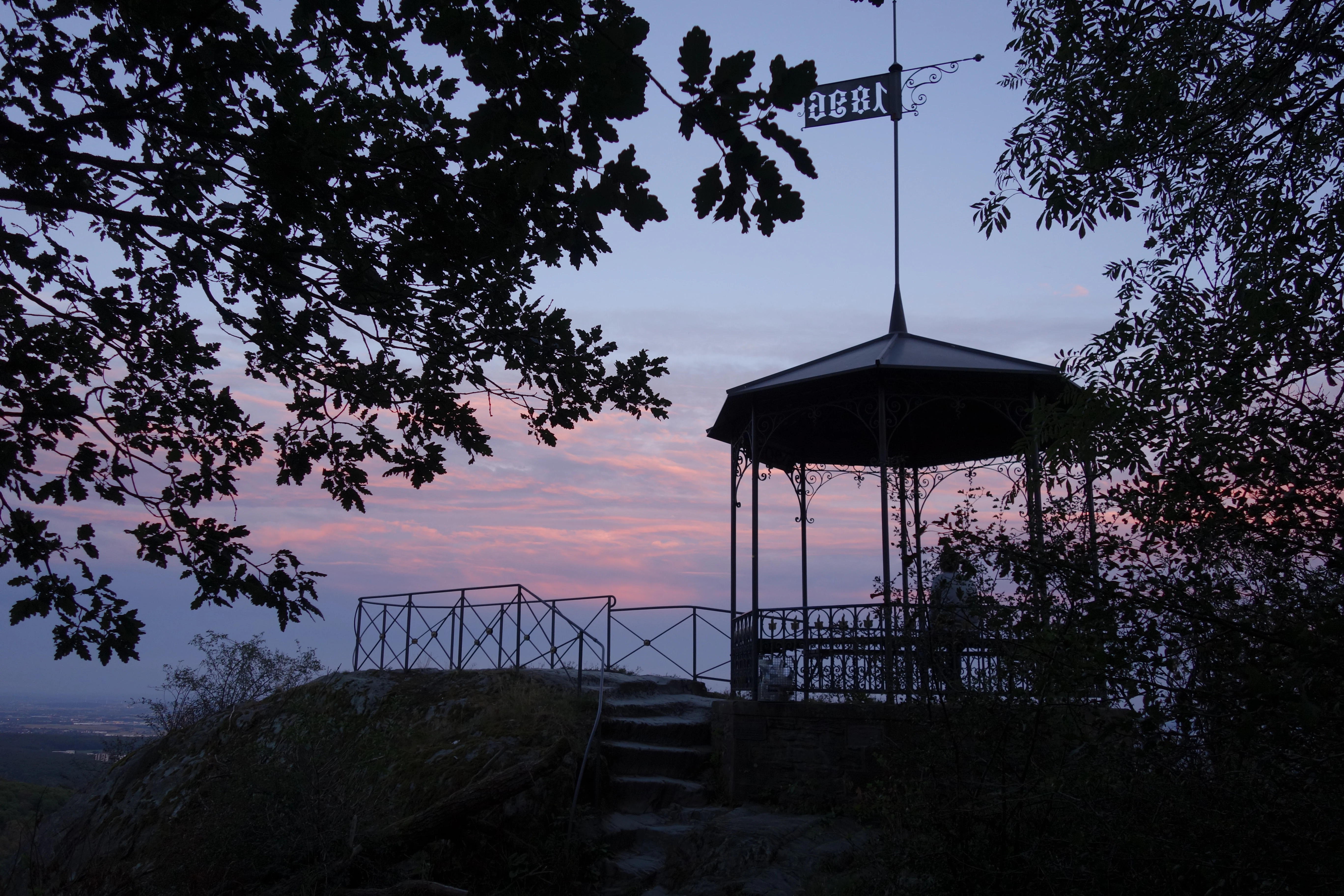 A silhouette of an ornate gazebo lookout structure with a metal roof and weather vane, set on a rocky outcrop against a colorful sunset sky, surrounded by tree branches.
