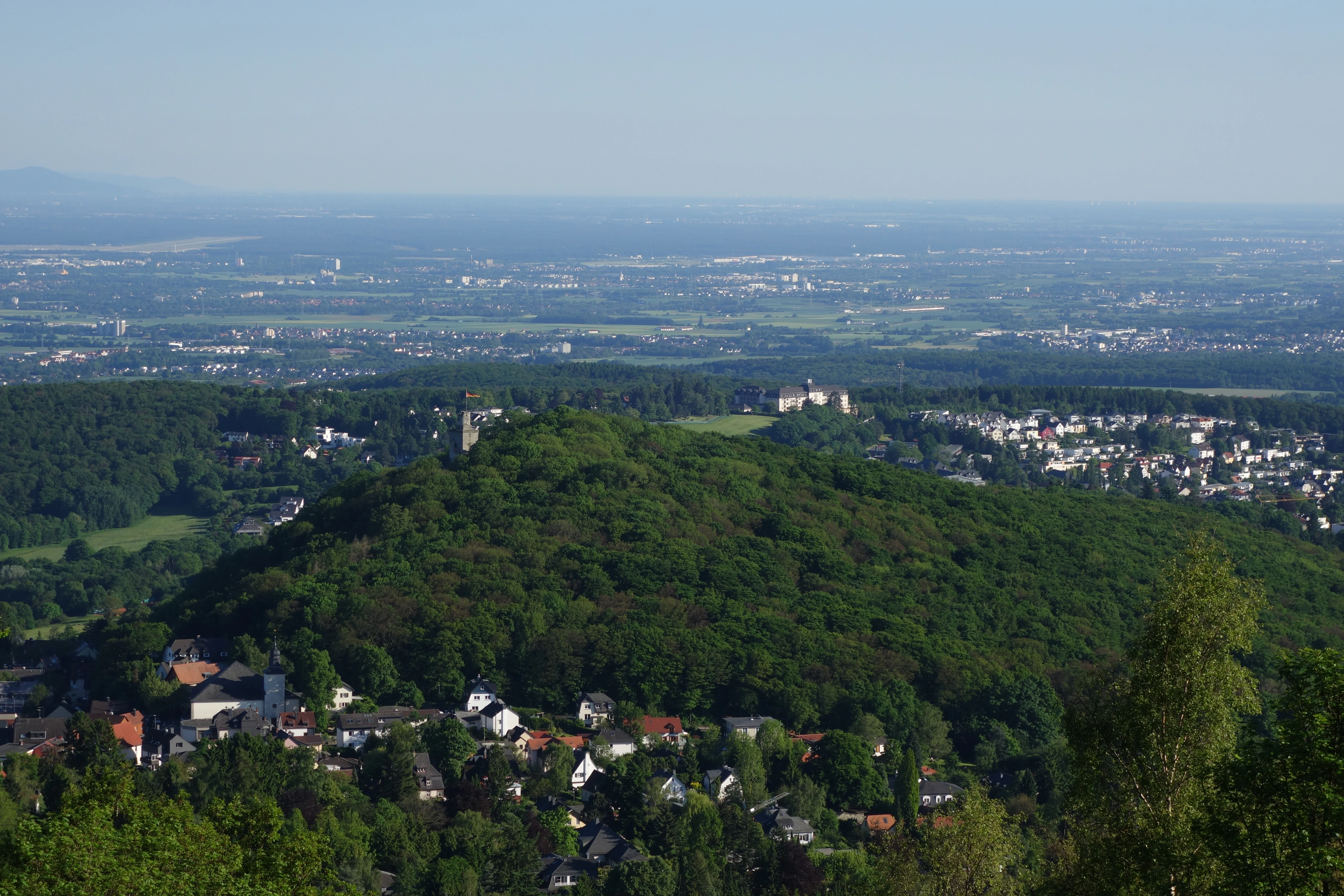 An expansive, elevated landscape view overlooking a forested hill, the ruins of Falkenstein Castle, and a residential area nestled in the valley, with a distant city skyline under a clear blue sky.