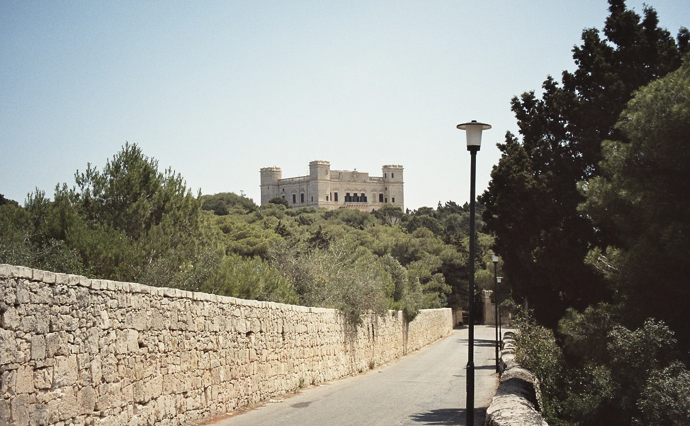 A stone road leads through a tree-lined area in Buskett Gardens toward the prominent stone Verdala Palace in the background.