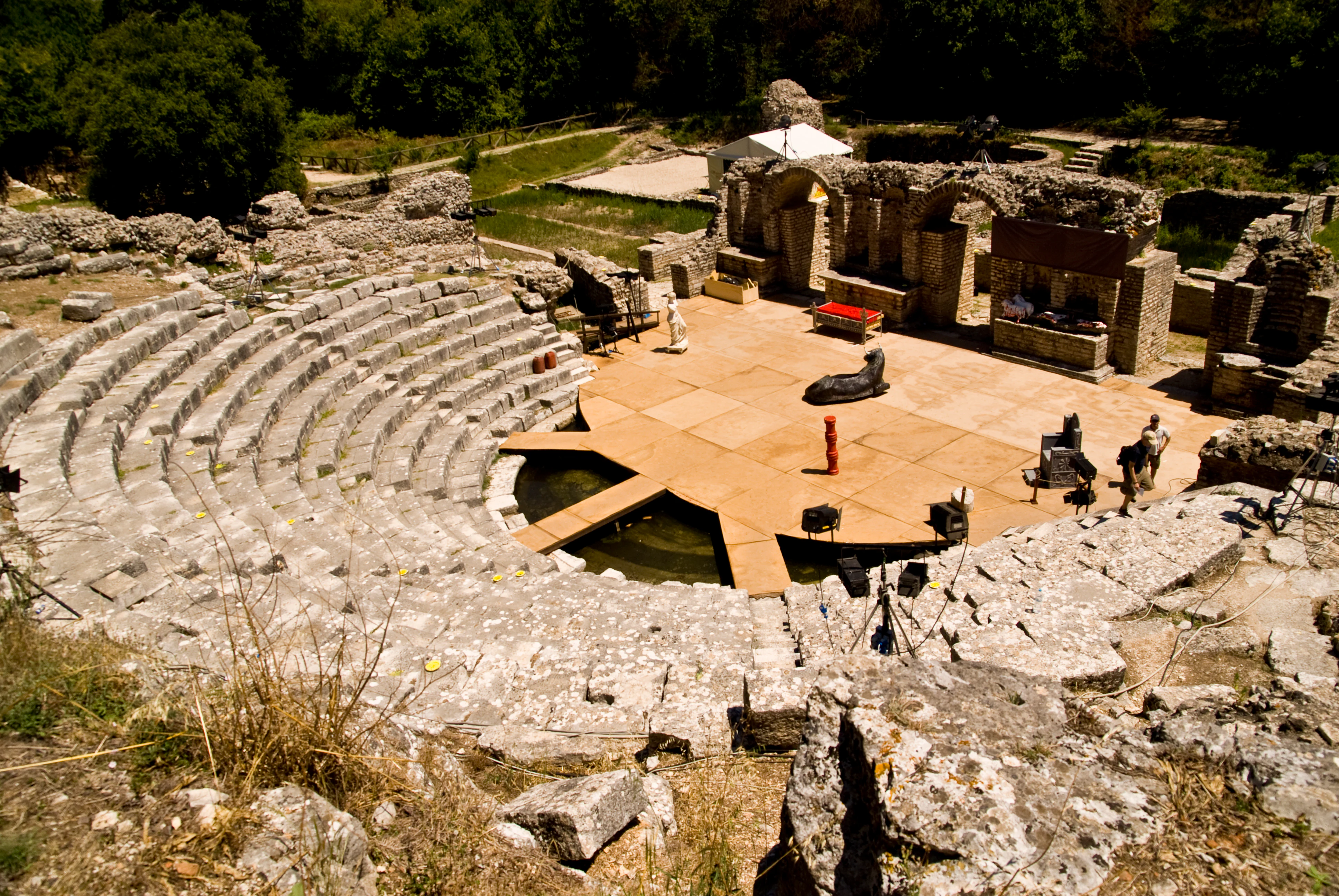 Stone amphitheater with tiered seating, surrounded by ruins of ancient structures and visitors, under a clear sky.