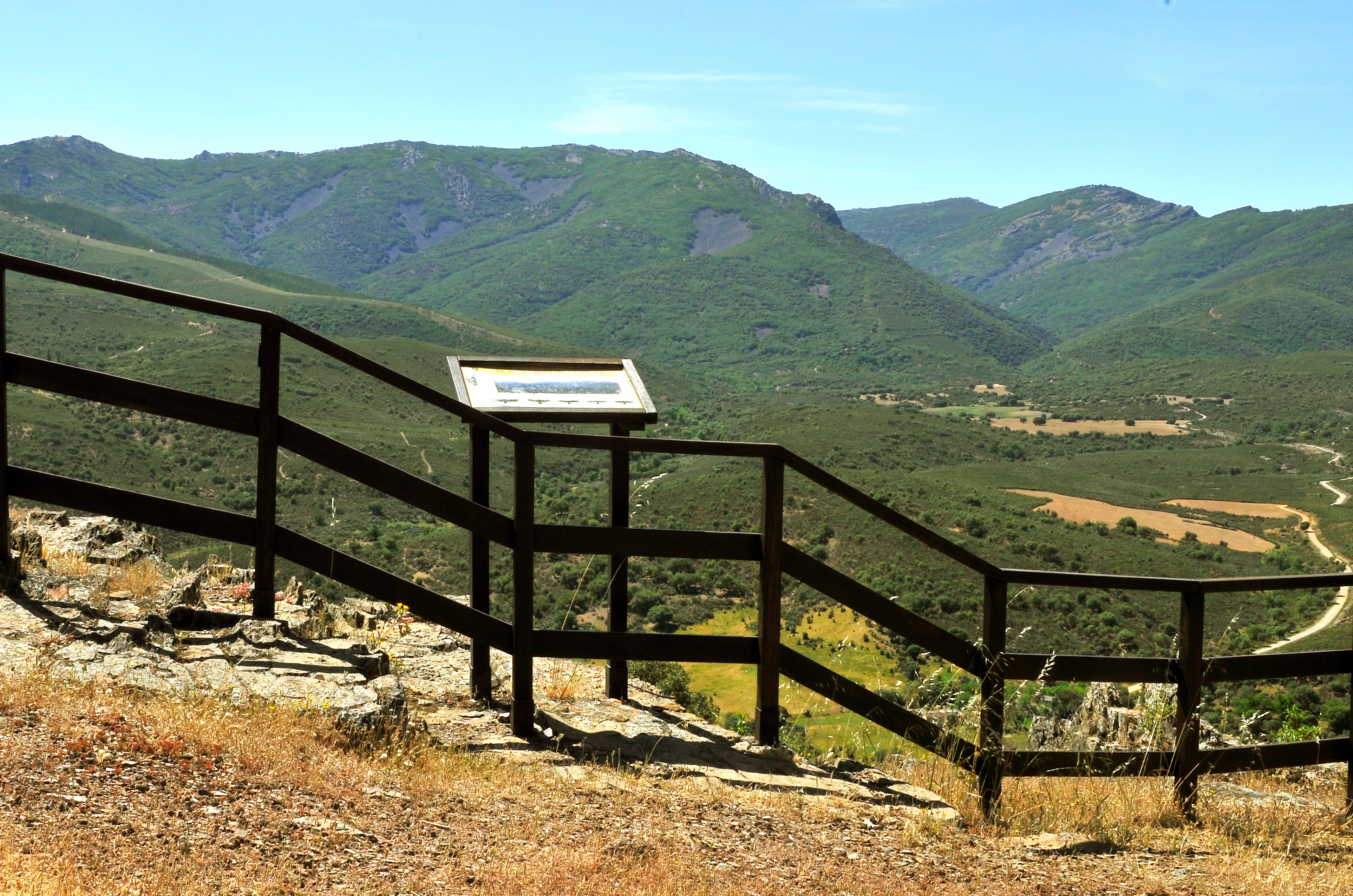 Wooden railing and sign at a viewpoint overlooking green mountains and valley