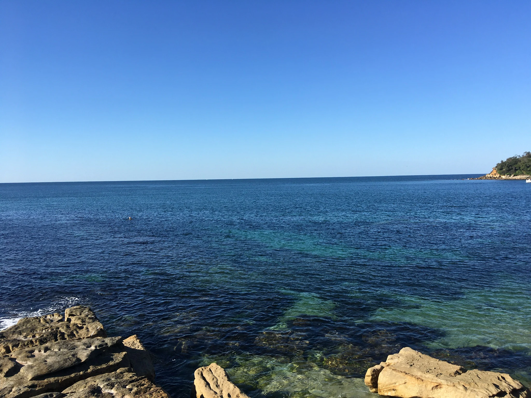 A wide view of a calm blue ocean bay with rocky foreground outcrops and a distant headland under a clear blue sky.