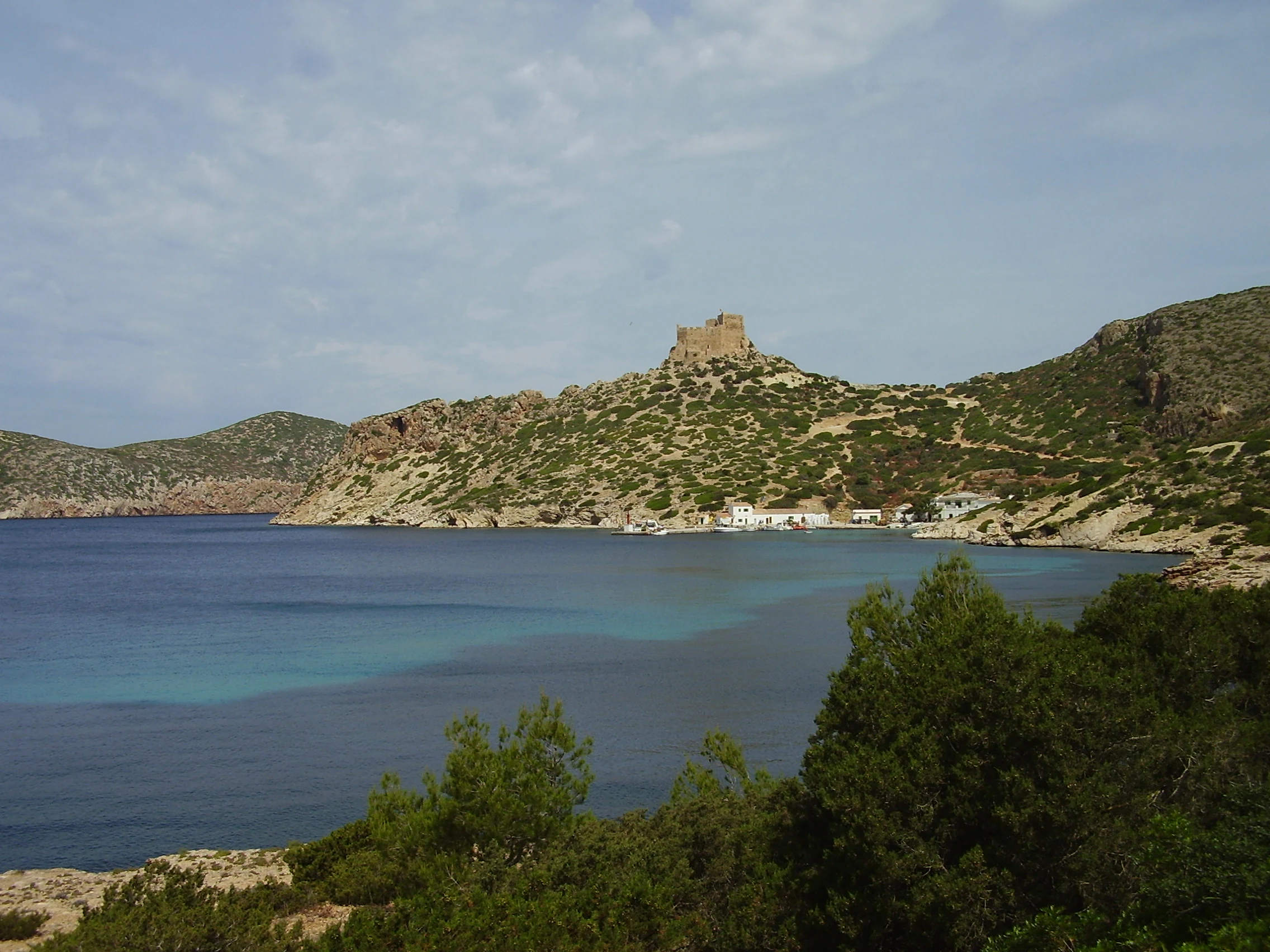 A coastal landscape view featuring a historic castle on a hilltop overlooking turquoise waters with rocky terrain and vegetation in the foreground