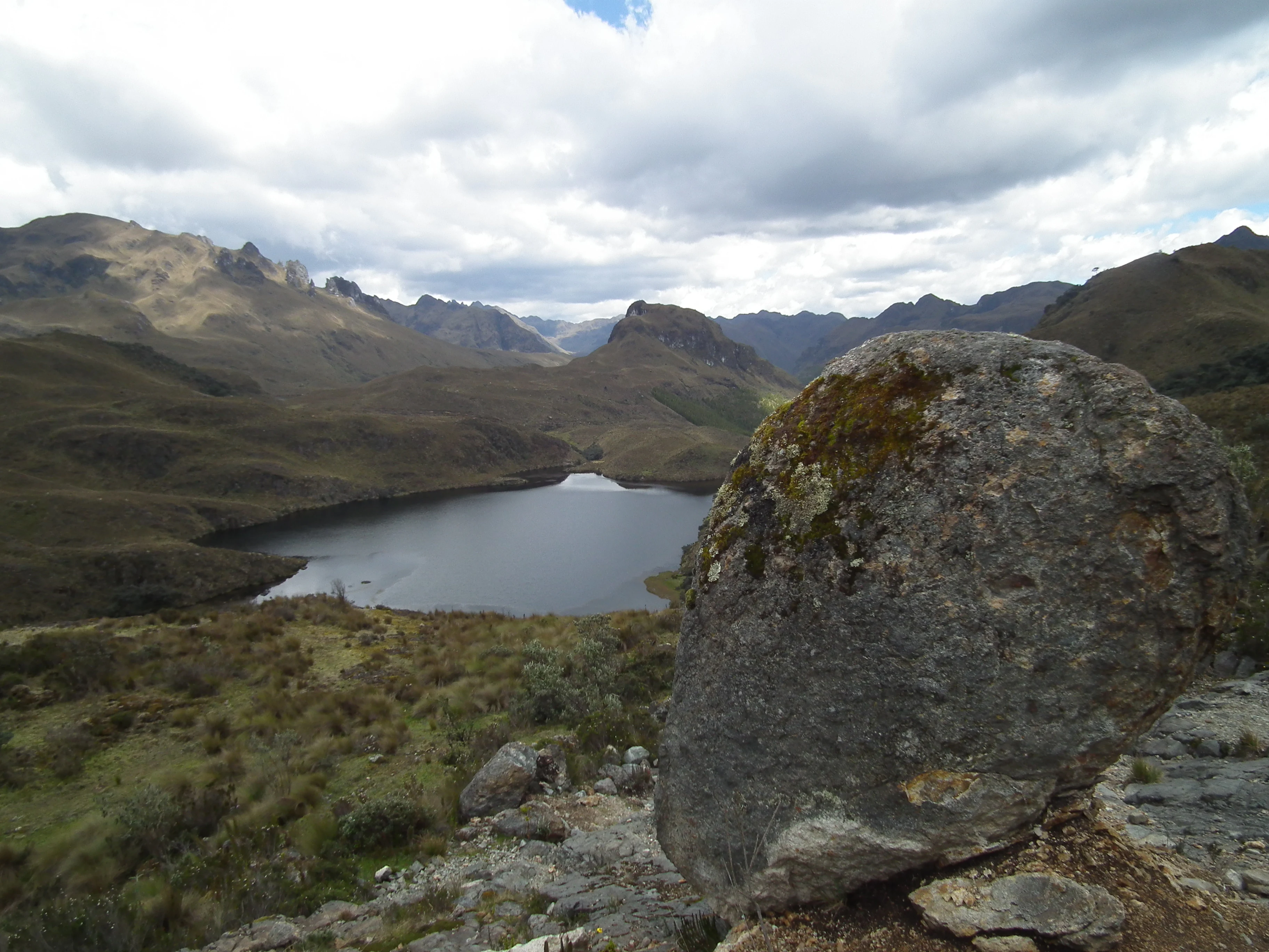 A wide, mountainous landscape featuring a dark glacial lake in the middle ground, with a large, lichen-covered boulder in the foreground and rolling, grassy hills under a cloudy sky.