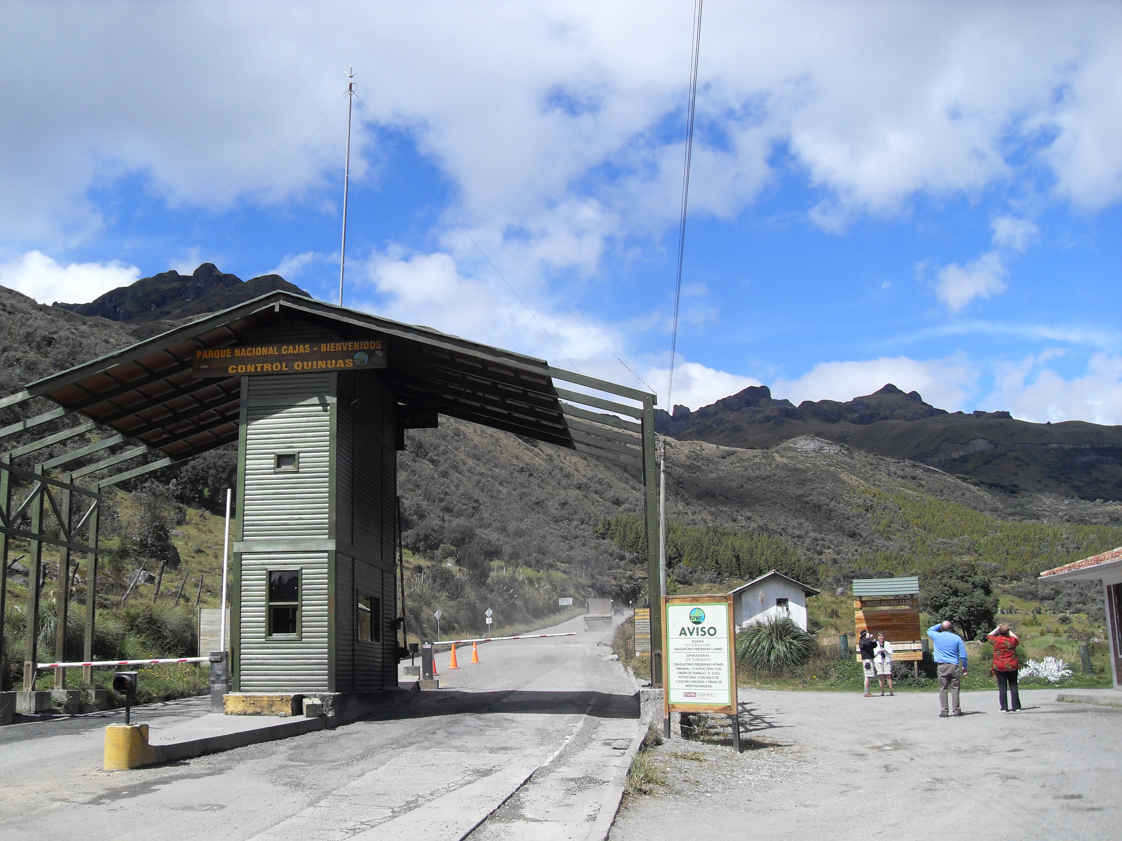 A park entrance gate structure made of green metal and wood with a small guard booth, set against a backdrop of Andean mountains.