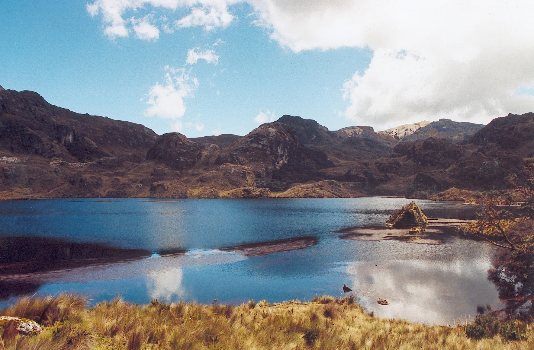 A wide scenic shot of a blue glacial lake surrounded by rolling, grass-covered Andean mountains under a sky with scattered clouds.
