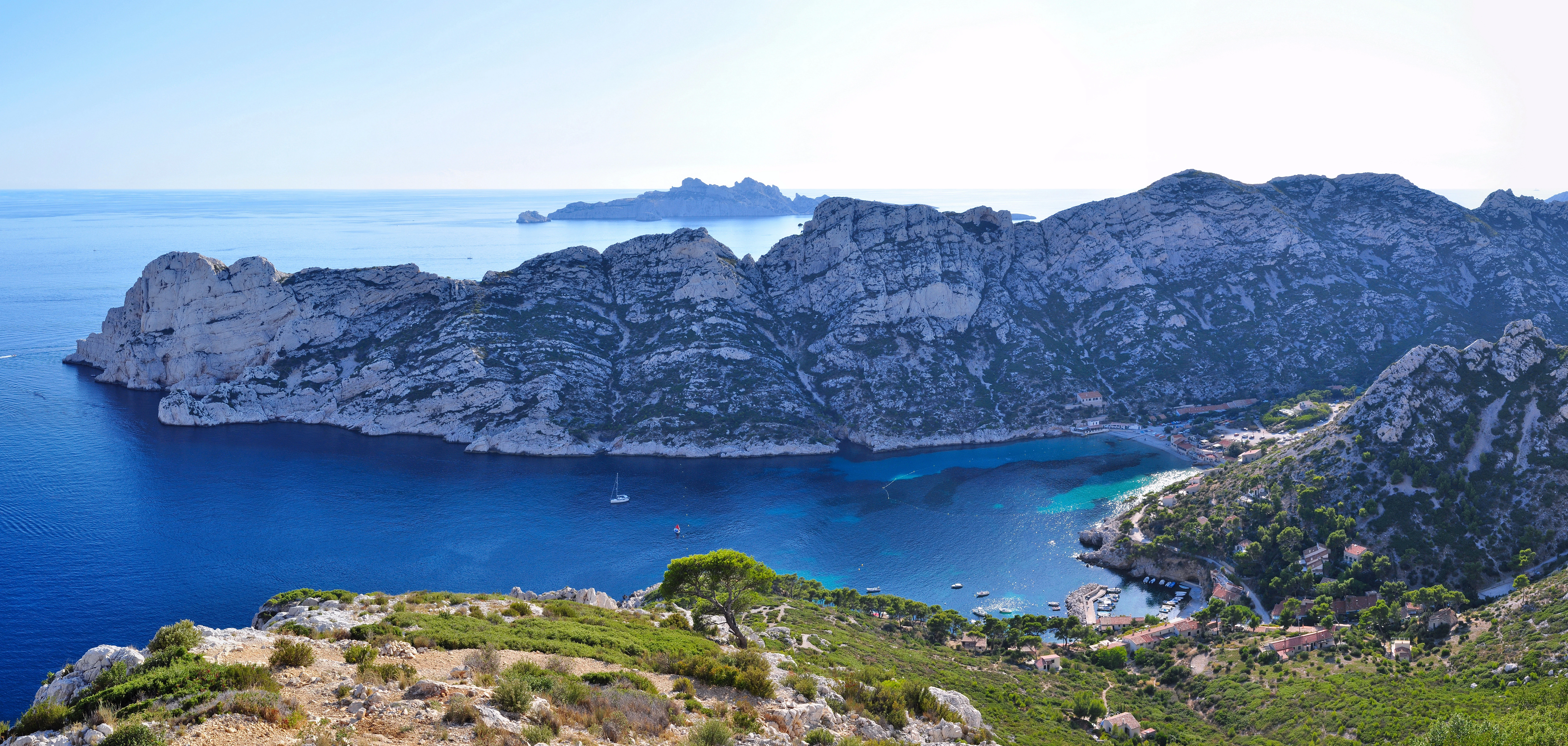 Wide view of a calanque with rocky cliffs, clear blue water, and green vegetation on a hillside under a bright sky