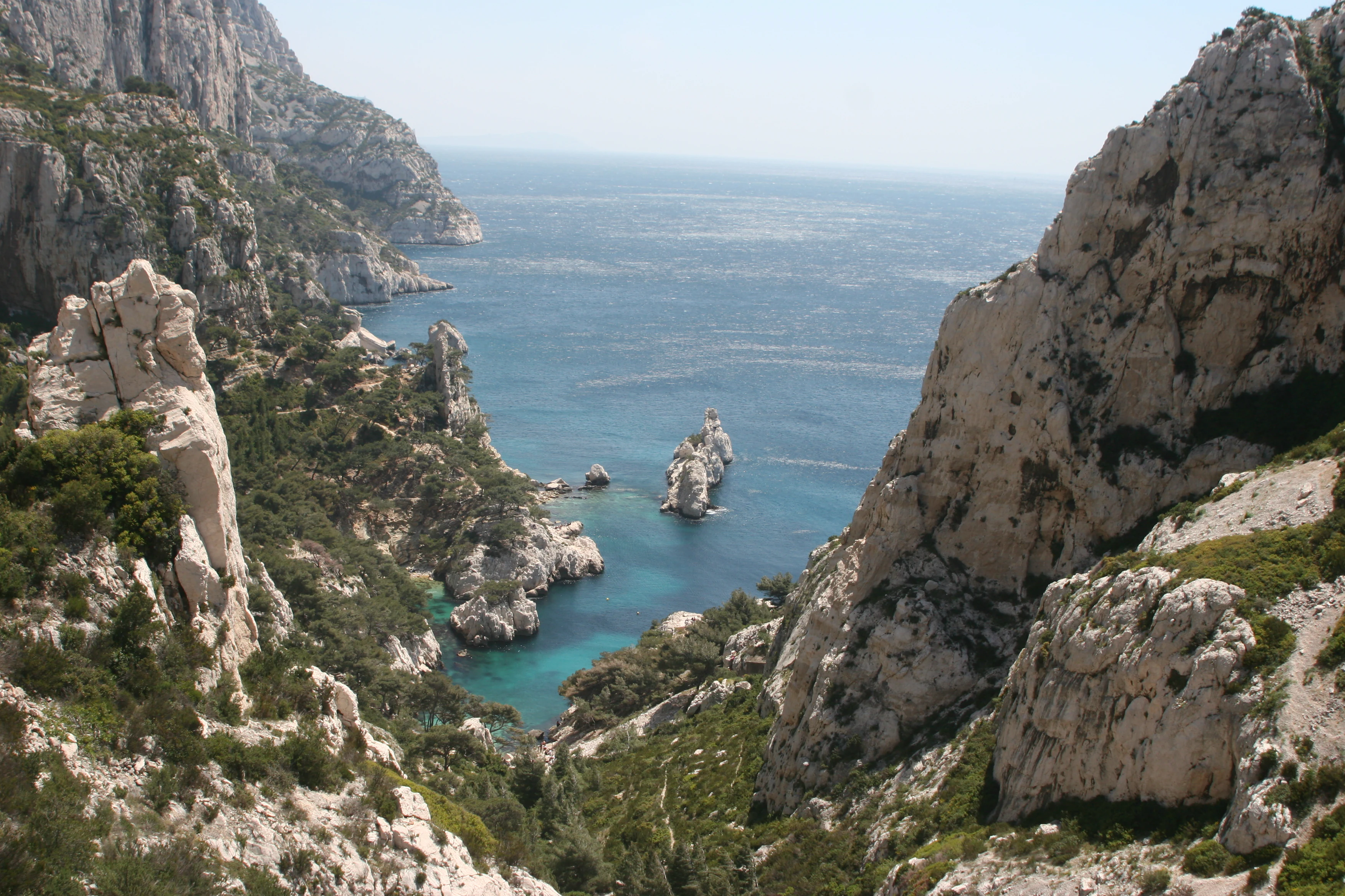 Rocky limestone cliffs forming a calanque with turquoise water, small rock formation in the sea, and green vegetation on slopes