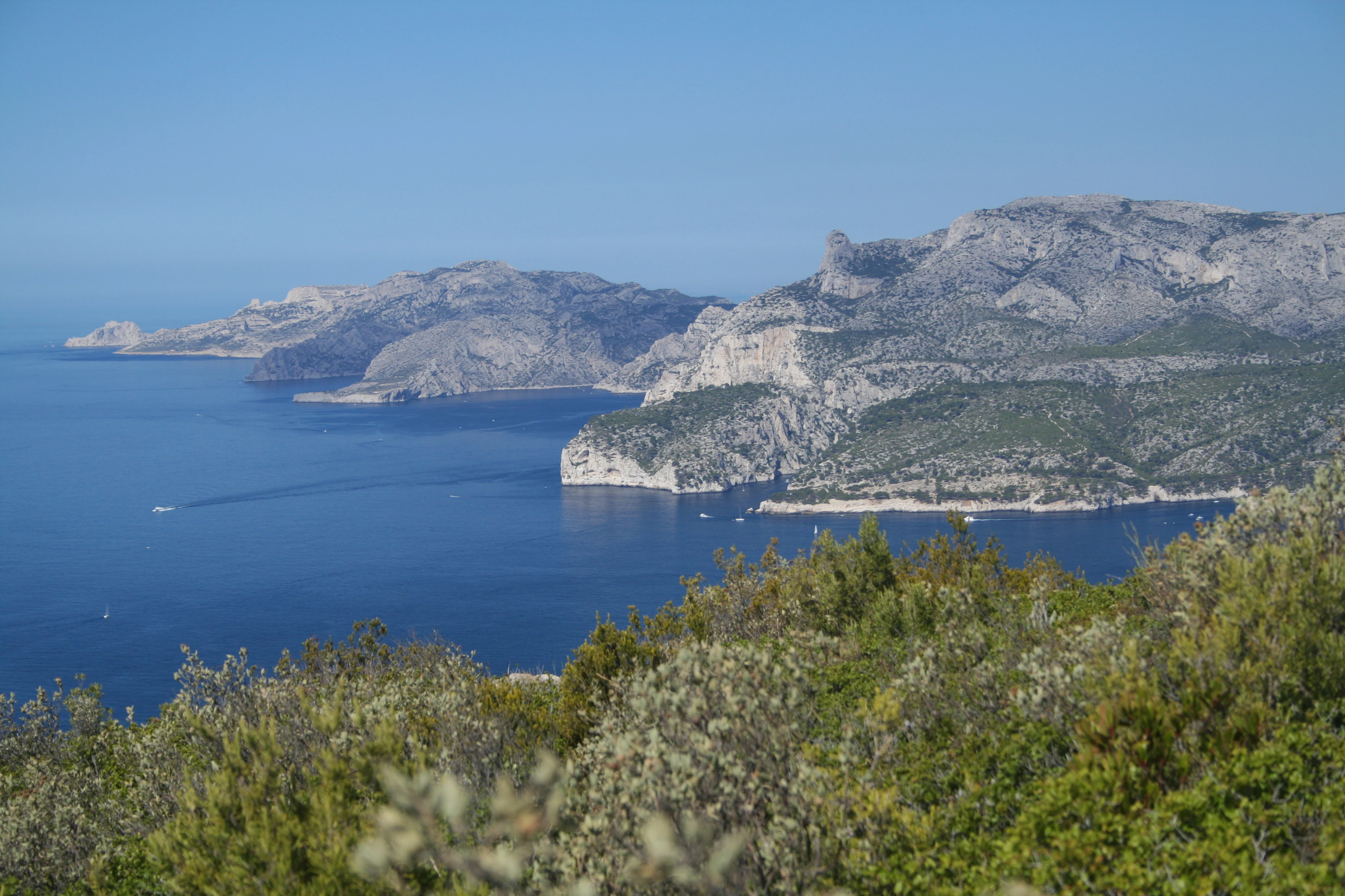 Blue sea, rocky coastal cliffs, and green vegetation with distant mountains under clear blue sky