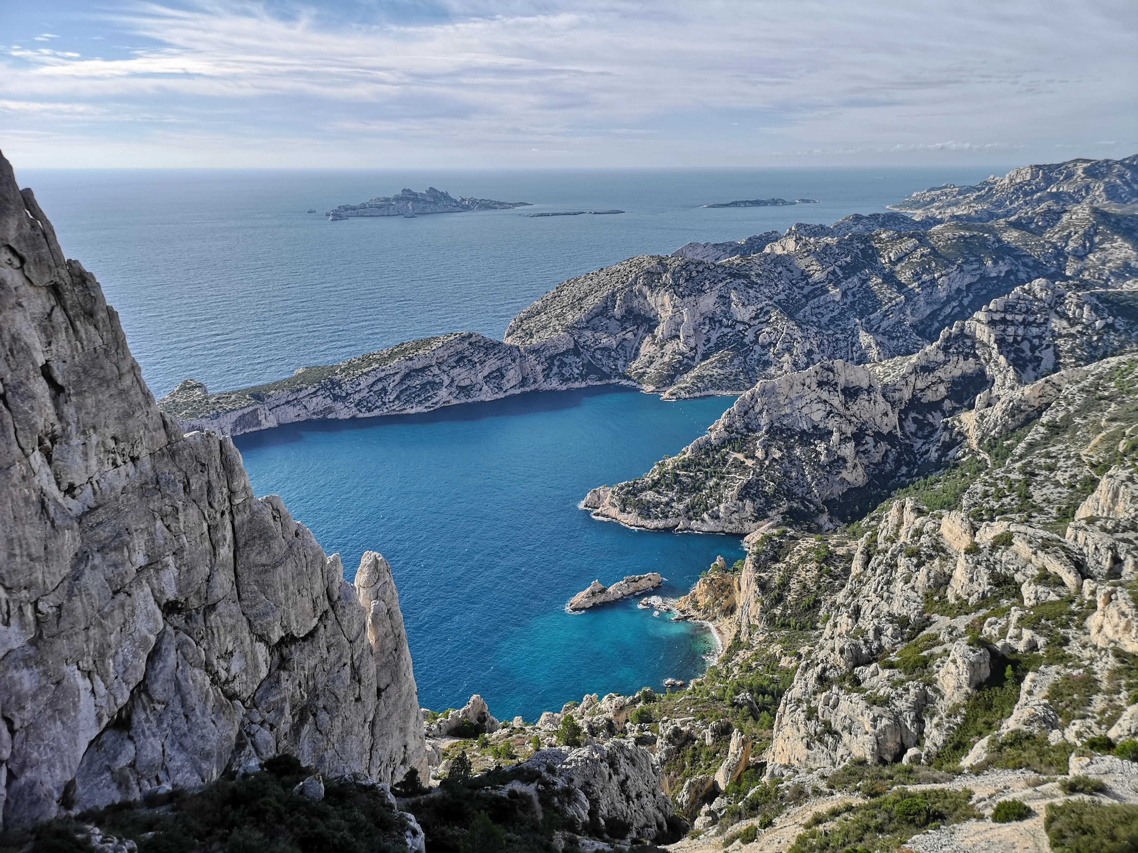 Rocky coastal cliffs overlooking turquoise water coves, distant islands, and mountainous terrain under a partly cloudy sky