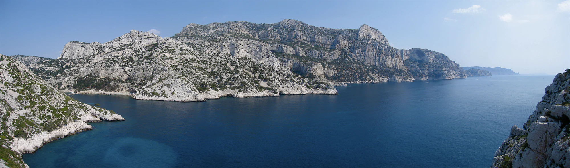 Wide-angle view of a rocky coastline with deep blue water, limestone cliffs, and distant mountains under a clear sky.