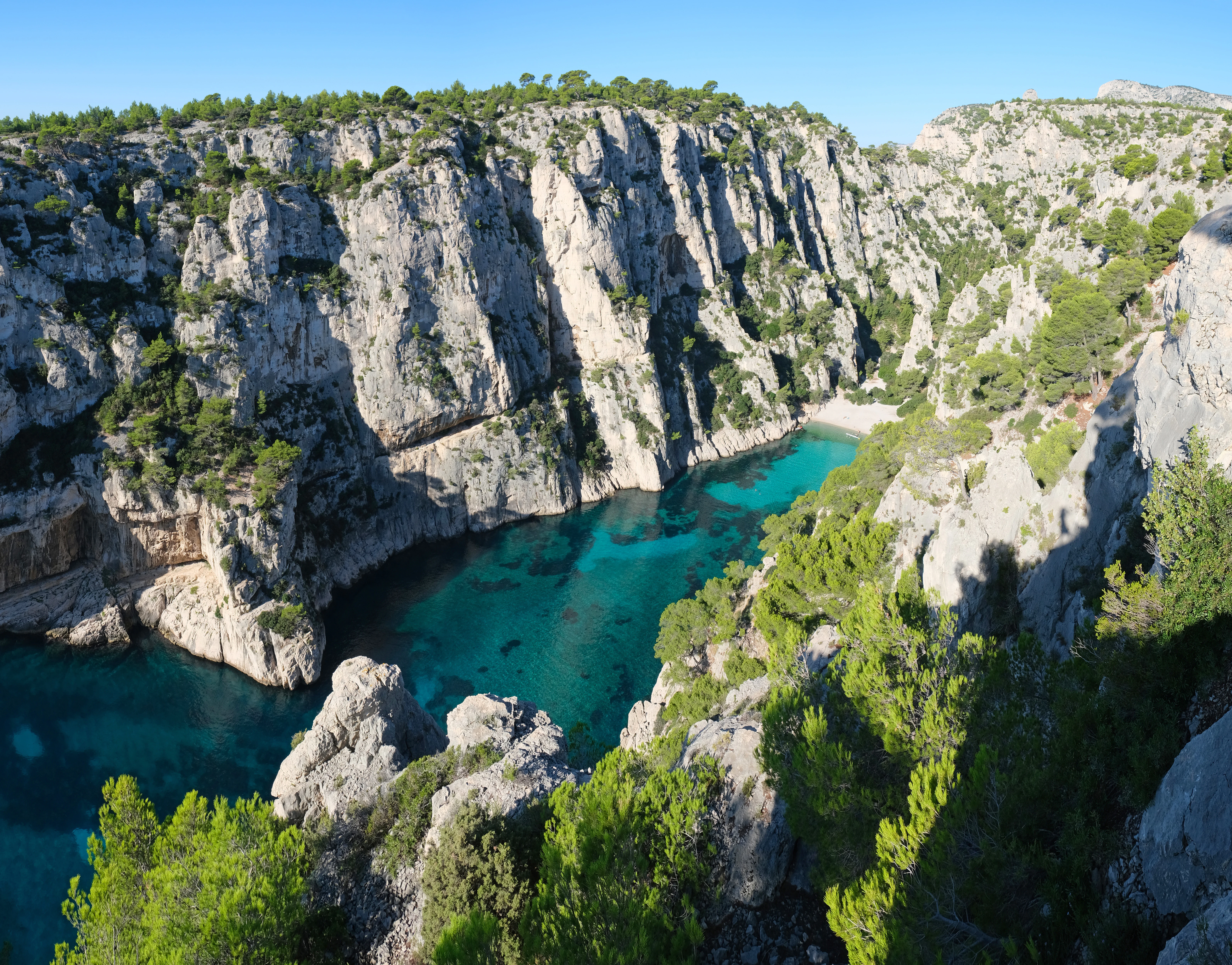 A dramatic limestone calanque with turquoise water surrounded by rocky cliffs and green vegetation under a clear blue sky.
