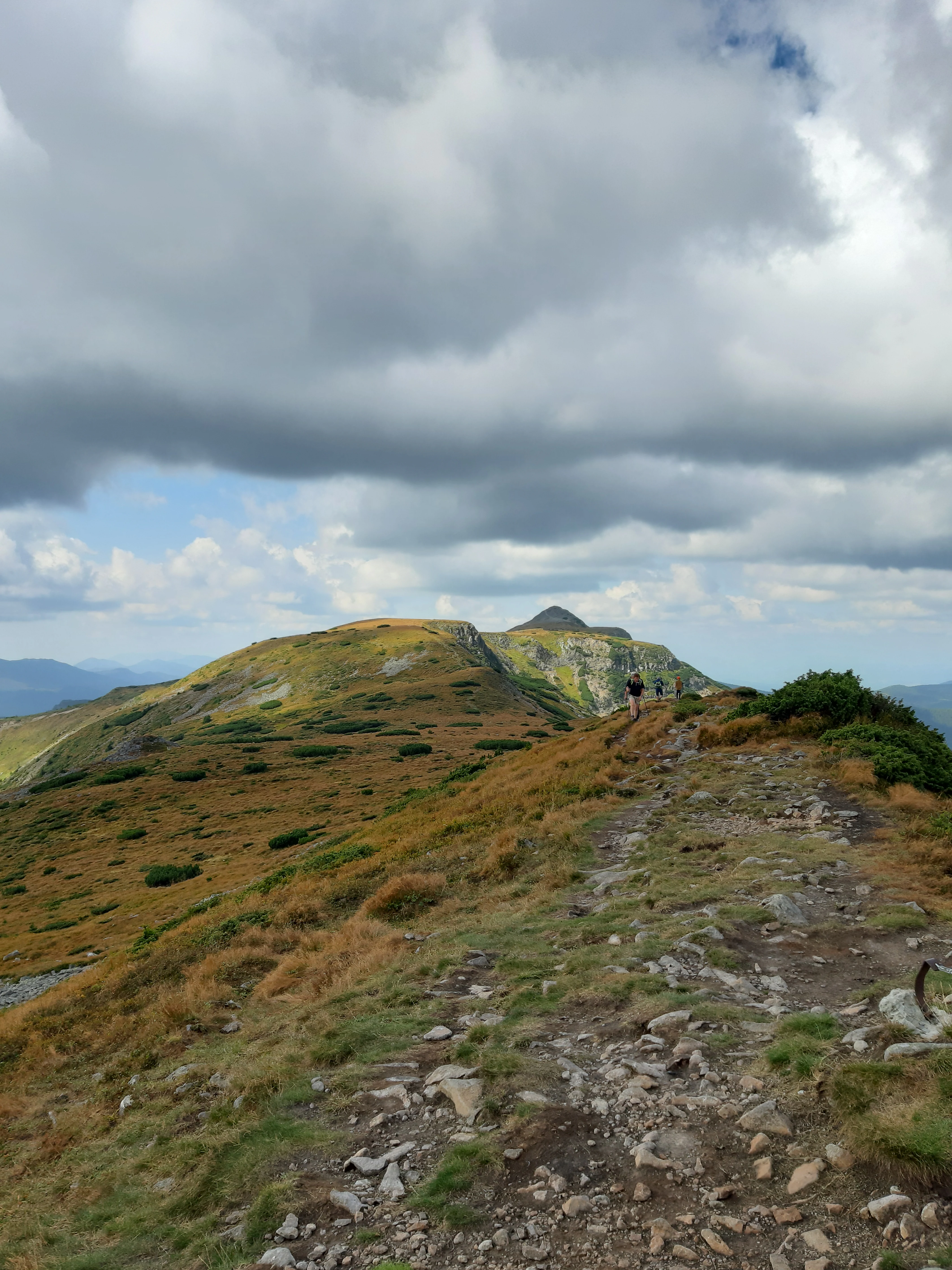 Rocky mountain trail ascending through grassy slopes with a hiker in the distance under a partly cloudy sky