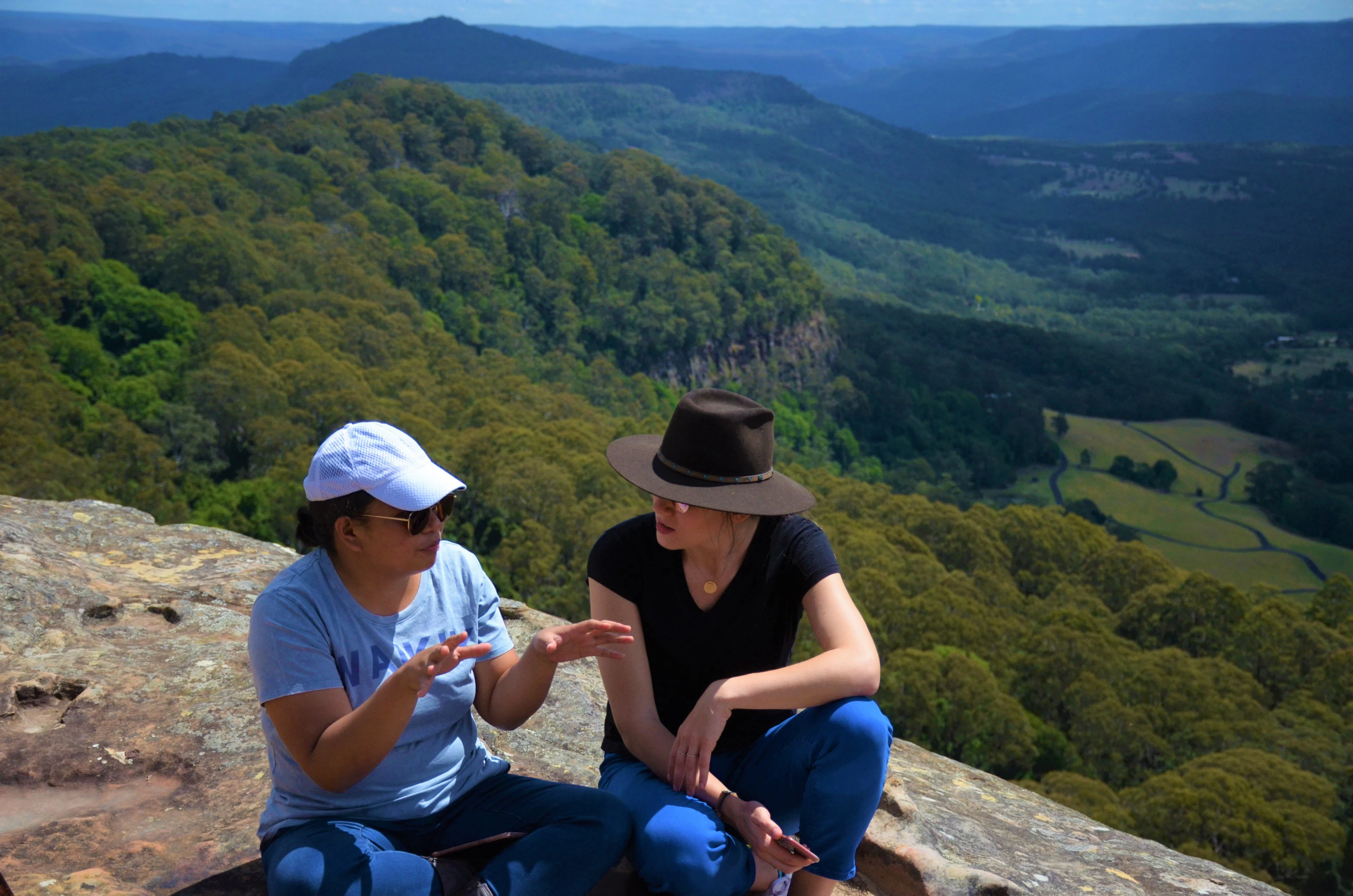 Two people sitting on a rocky cliff edge, talking while overlooking a vast, lush green forest valley and distant ridgelines in the Cambewarra Range.