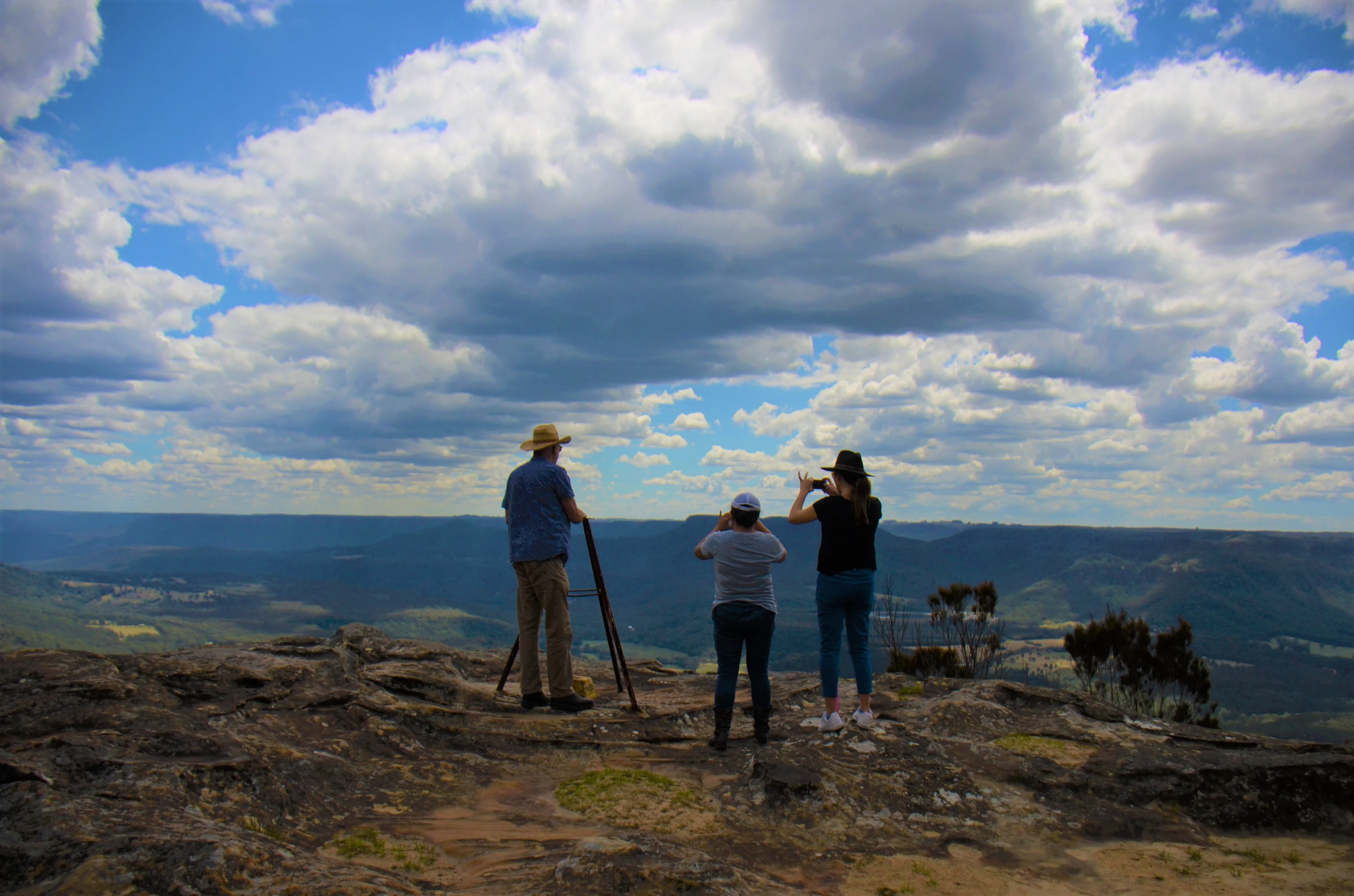 Three people stand on a rocky outcrop looking out over a wide valley under a blue sky with scattered clouds.