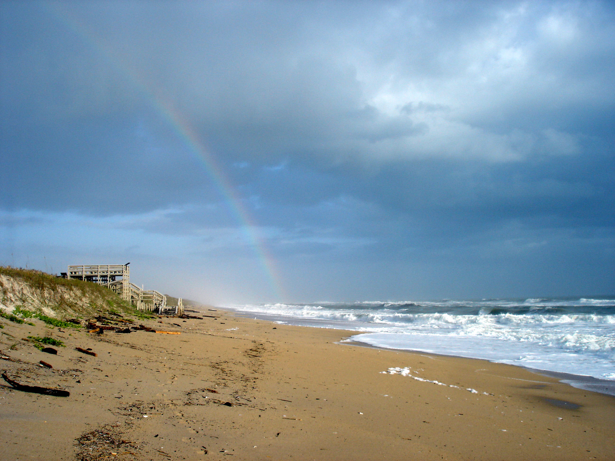 A wide view of a sandy beach coastline with ocean waves and a faint rainbow in a cloudy sky. A wooden boardwalk ramp is visible on the dune in the foreground.