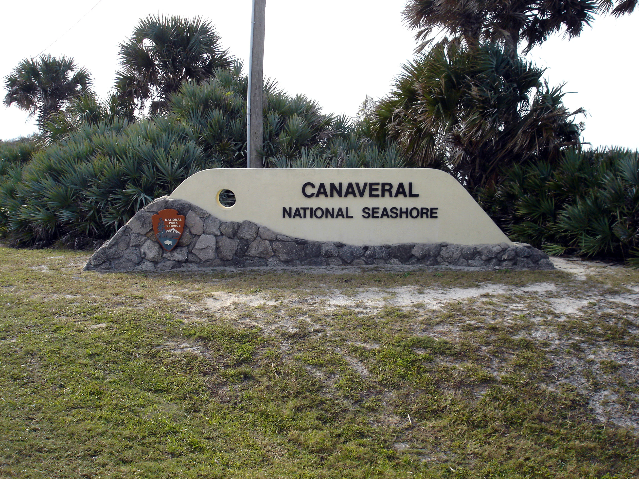A beige-colored sign with the text Canaveral National Seashore resting on a base of stones, surrounded by palm trees and coastal vegetation.